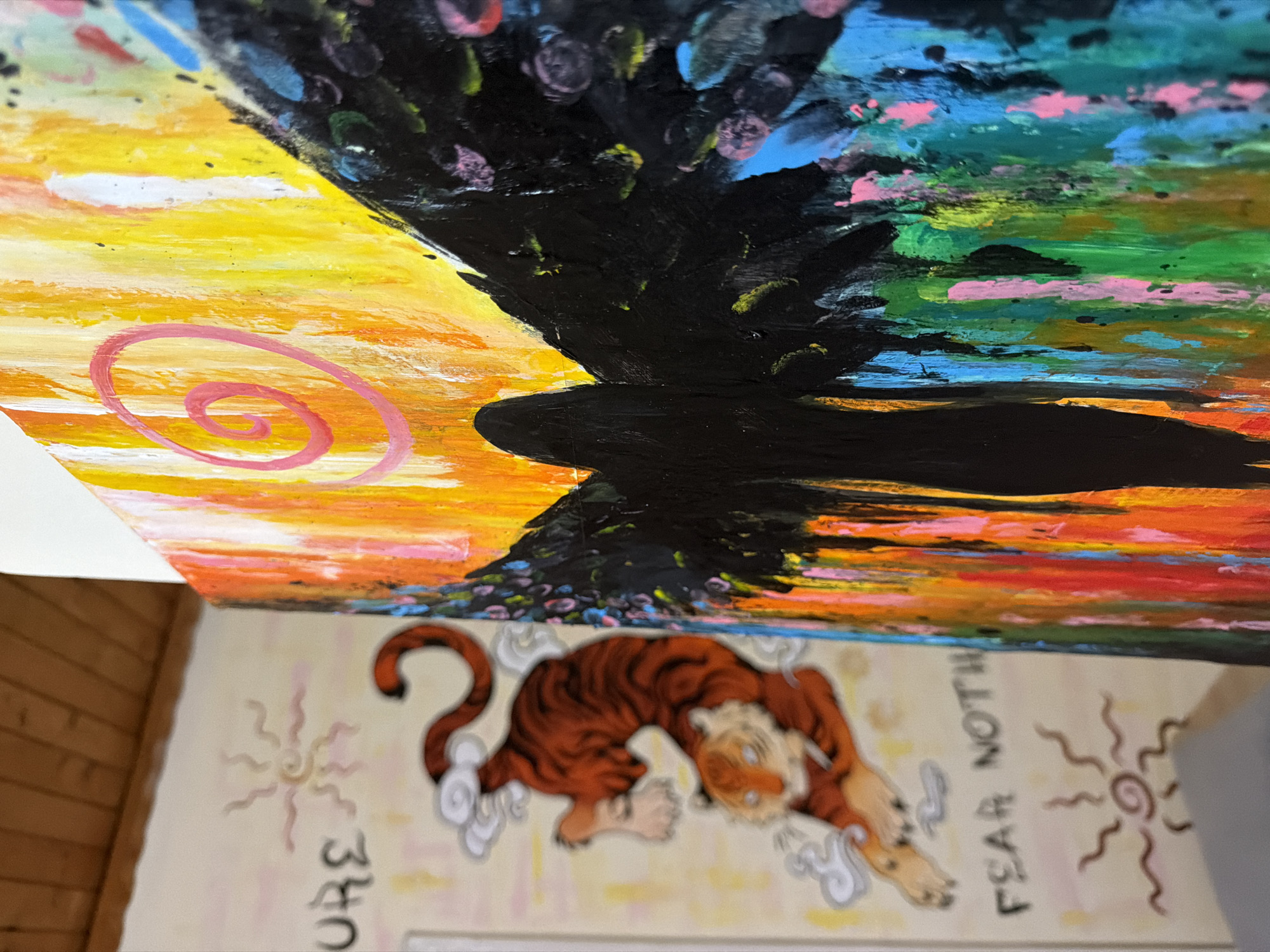 The image shows a close-up of two paintings in Svilengrad, Bulgaria. On the left, a painting on a light-colored wall depicts a stylized orange tiger in a jumping pose, surrounded by white clouds. Above the tiger, the word "URE" is partially visible, and below it, the phrase "FEAR NOT" is written in black. A small, sun-like symbol with wavy rays is also present in the lower section of this painting. To the right, a second, more abstract painting is shown. This painting features a dark silhouette of a figure with large wings, rendered in black. The background behind the figure is a vibrant blend of colors, including yellows, oranges, pinks, greens, blues, and black, with visible brushstrokes and paint drips. A pink spiral is prominently displayed in the upper right section of this second painting. The wooden ceiling of a room is visible in the top left corner of the image.