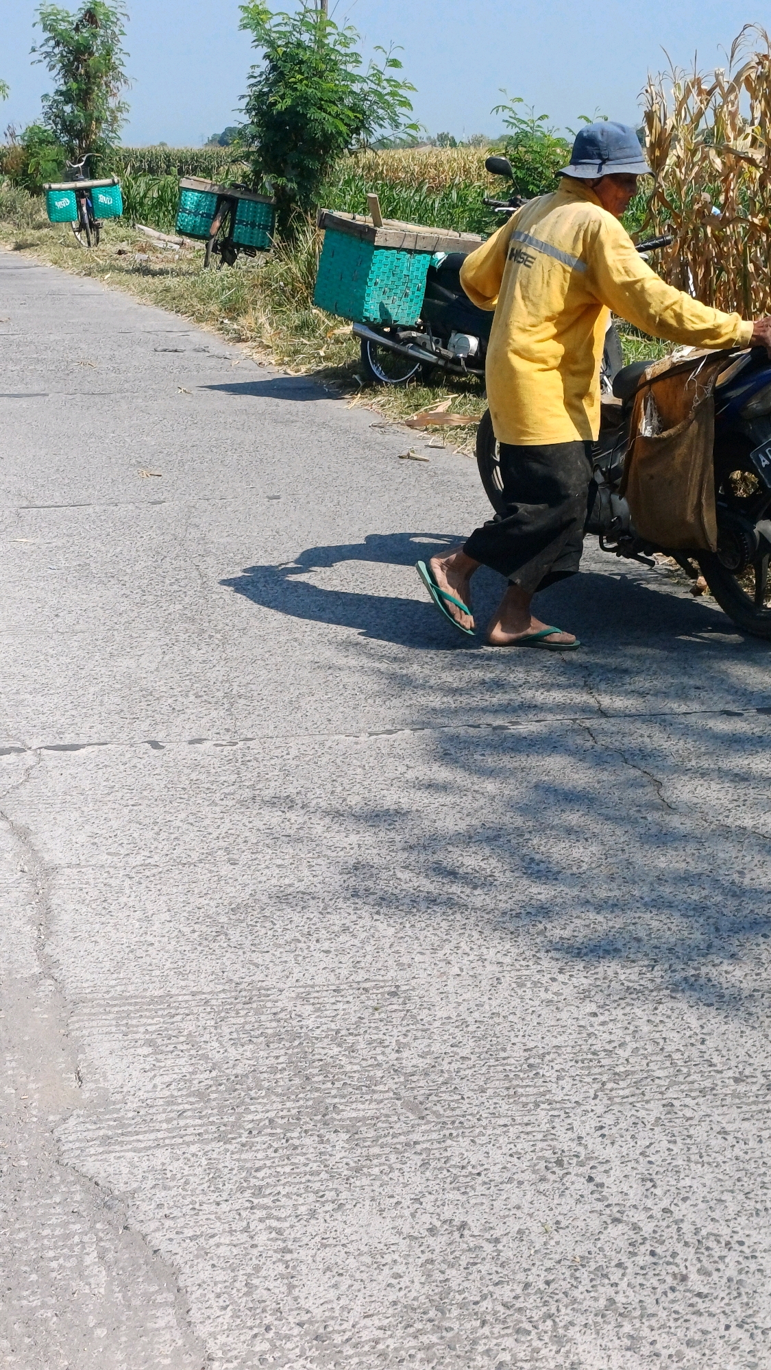 A person wearing a yellow long-sleeved shirt, black pants, and a blue hat walks beside a motorcycle parked on a paved road. The person is pushing or leaning on the motorcycle. The motorcycle has a brown woven bag attached to the rear. In the background, there are several bicycles with green woven baskets, and a field of dry corn stalks. The scene appears to be in a rural or semi-rural area.
