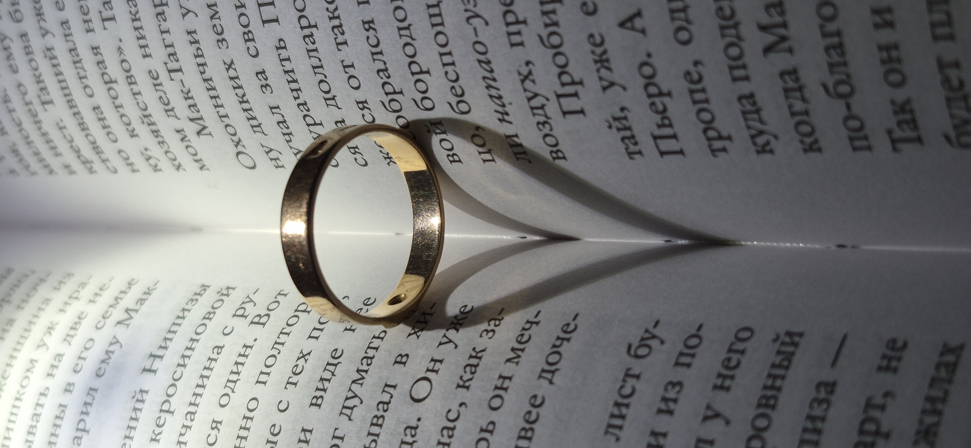 The image displays an open book with a gold ring placed on its pages. The book is positioned vertically, with the spine in the center, creating a heart-shaped shadow formed by the folded pages and the ring. The text on the pages is in Russian. The lighting suggests an indoor setting, possibly a home or a study, with a single light source casting dramatic shadows. There are no people or animals visible in the image. The image does not contain any information or context to suggest a location in Ussurijsk, Russia.