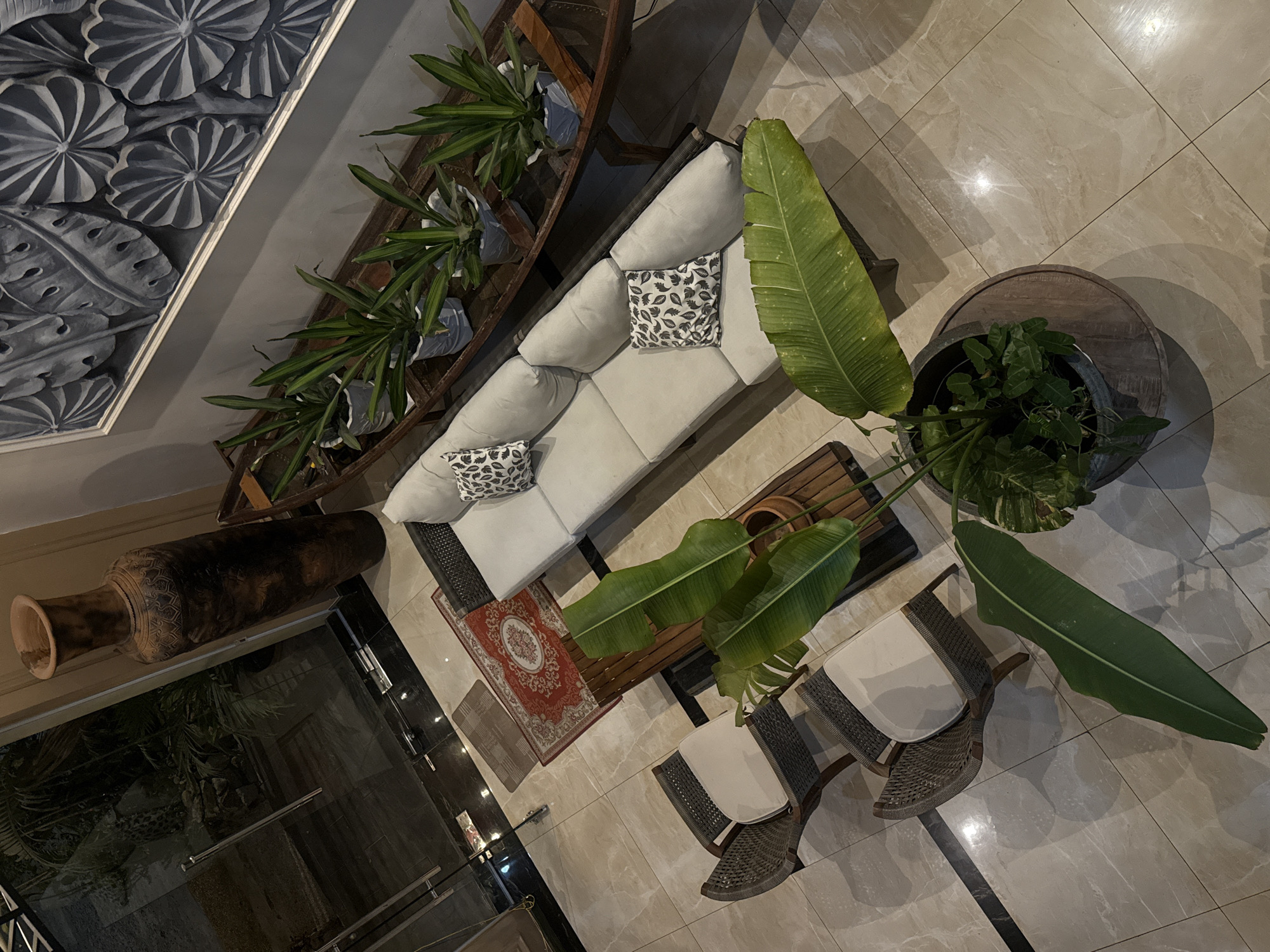 This high-angle, full-shot photograph captures a well-appointed indoor space, possibly a lobby or lounge area in Bac Nha Trang Ward, Vietnam. The scene is dominated by a large, light-colored sectional sofa adorned with two patterned throw pillows. In front of the sofa sits a low wooden coffee table with a dark wood plank construction, topped with a large potted plant featuring broad green leaves. To the left of the sofa, two wicker armchairs with light-colored cushions are arranged.
Behind the sofa, a long, narrow wooden boat-like structure serves as a planter for several potted green plants, adding a touch of nature to the indoor setting. Above this arrangement, a large textured artwork featuring stylized leaf motifs is mounted on the wall.
To the left of the main seating area, large glass doors offer a glimpse of an outdoor space with greenery, suggesting the proximity to nature or a garden. A tall, ornate dark brown vase stands near the glass doors, adding a decorative element. A small, intricately patterned red rug is placed on the beige tiled floor between the glass doors and the sofa. The floor itself is composed of large, light-colored tiles with subtle veining, reflecting ambient light. The overall impression is one of a comfortable and stylish waiting or relaxation area.