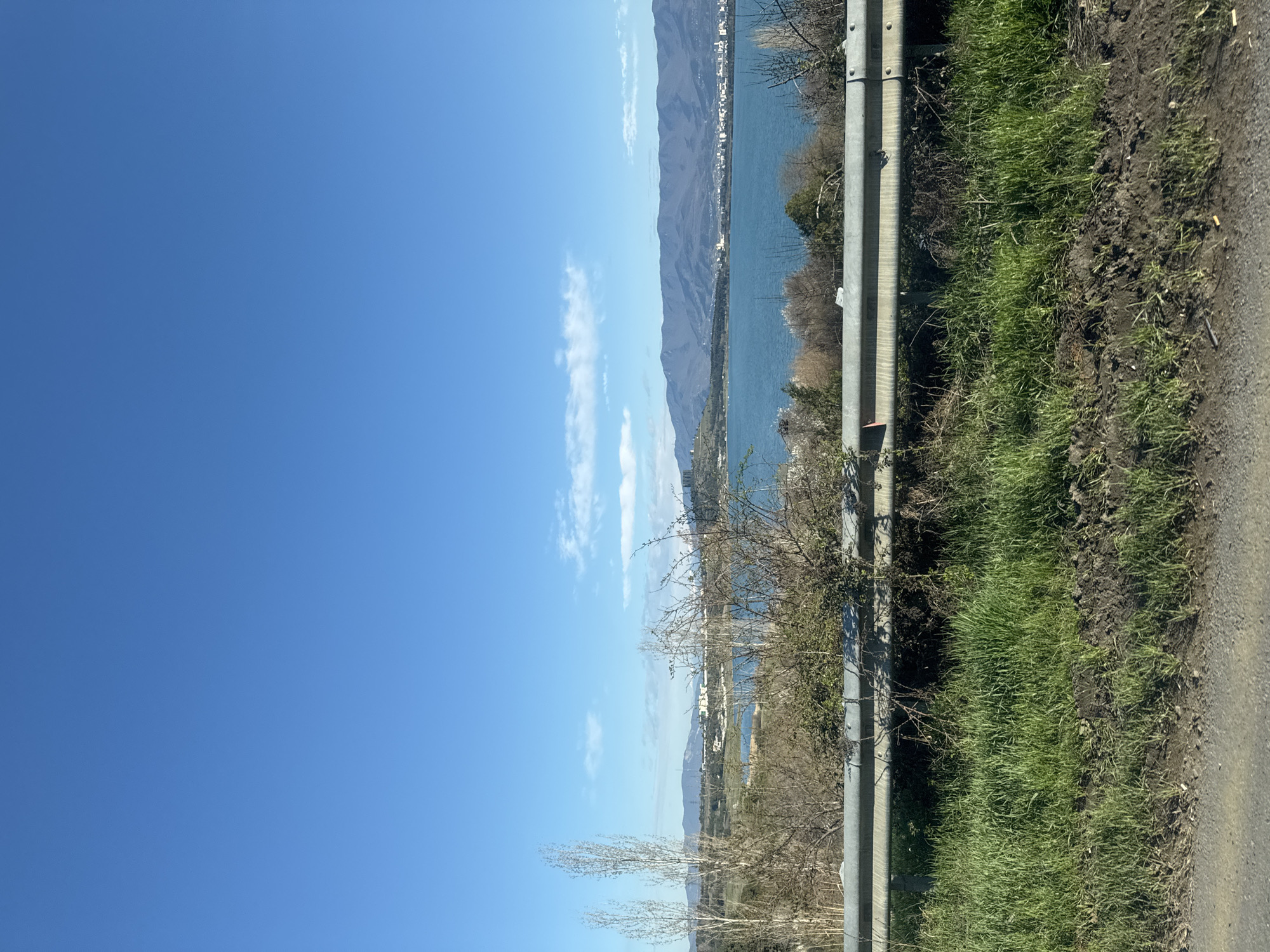 The image depicts a landscape taken from the side of a road, potentially near Tbilisi, Georgia, as the terrain and architecture in the background resemble that of the city and its surroundings. A guardrail is visible in the foreground, separating the road from a grassy area that leads down to a body of water. Beyond the water are mountains, and the sky is mostly clear with a few scattered clouds. Buildings and urban structures are visible along the water's edge and nestled against the mountains. The presence of bare trees suggests it might be late winter or early spring.