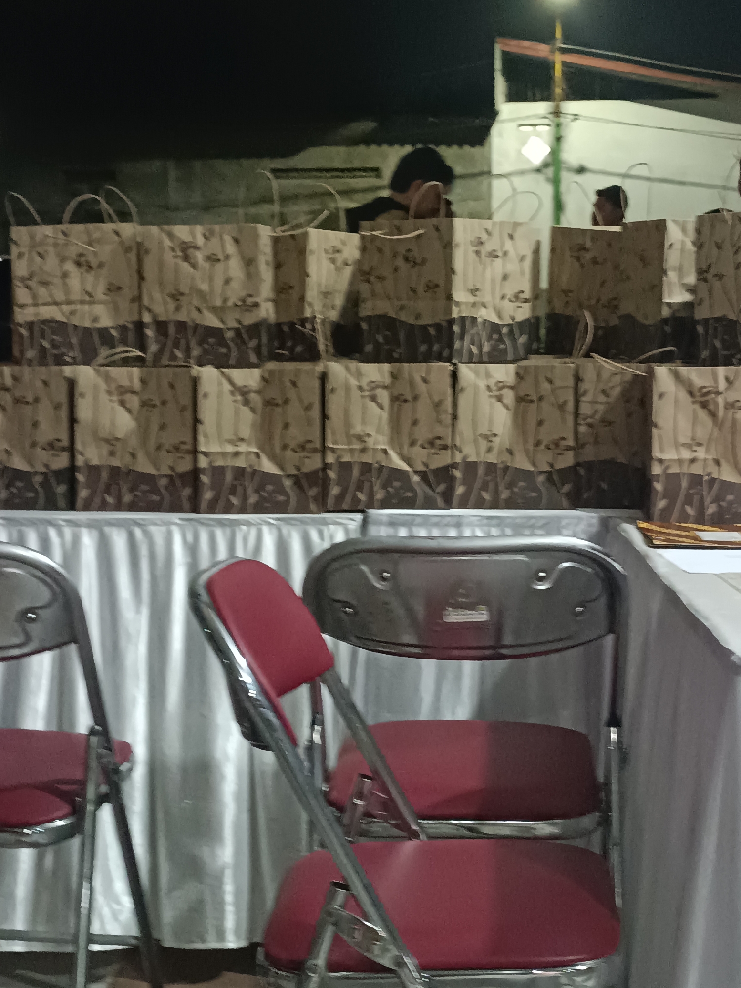 The image shows a row of paper gift bags with a floral pattern, stacked on top of each other. In the foreground, there are several red and silver folding chairs. Behind the chairs and the bags, a white tablecloth covers a surface. In the background, there are two individuals standing, and a building with a light pole and some wiring. The scene appears to be set up for an event, possibly in Condongcatur, Indonesia.