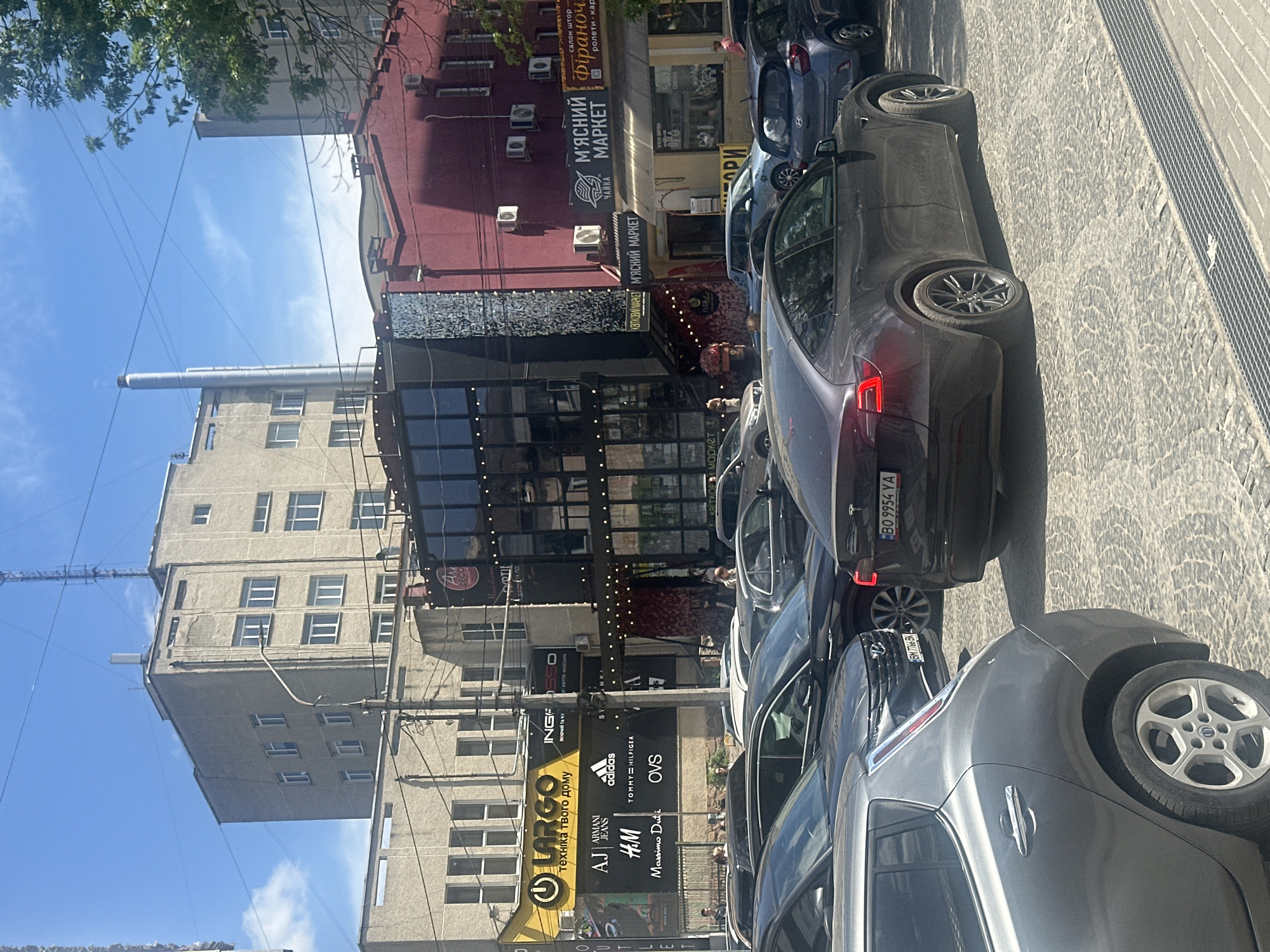 The image shows a street scene in Ternopil, Ukraine, with several parked cars along a cobblestone street. A multi-story building with a gray facade is visible, as well as a building with a red exterior that contains the words "М'ЯСНИЙ МАРКЕТ" (Meat Market) in Ukrainian on a sign. In front of this building is a storefront with the name Largo, which has many well-known brand names listed.
A tall industrial chimney is visible in the background against a blue sky with scattered clouds. There are power lines and cables in the foreground and background. The focus is on the cars and buildings lining the street, indicating an urban or commercial environment. The angle of the image suggests it was taken from street level, possibly by a pedestrian or someone in a vehicle.
