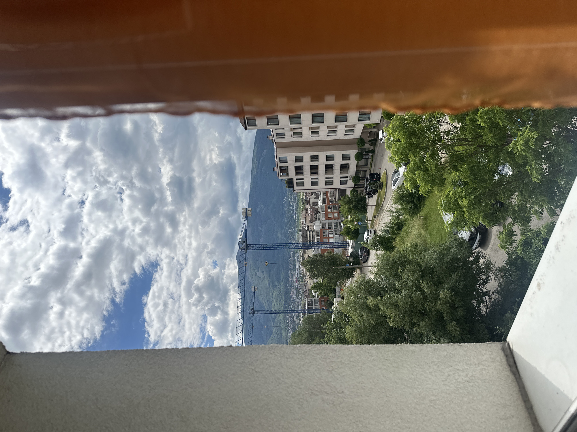 A wide-angle, eye-level outdoor shot captures a view of Sofia, Bulgaria, framed by a textured white wall on the left and a blurry orange object on the right. The middle ground features a cityscape with buildings, trees, and a road with parked cars. In the background, two large blue construction cranes stand out against a backdrop of lush green mountains under a cloudy sky. The scene appears to be taken from a high vantage point, possibly a window or balcony, overlooking a residential and construction area in Sofia.