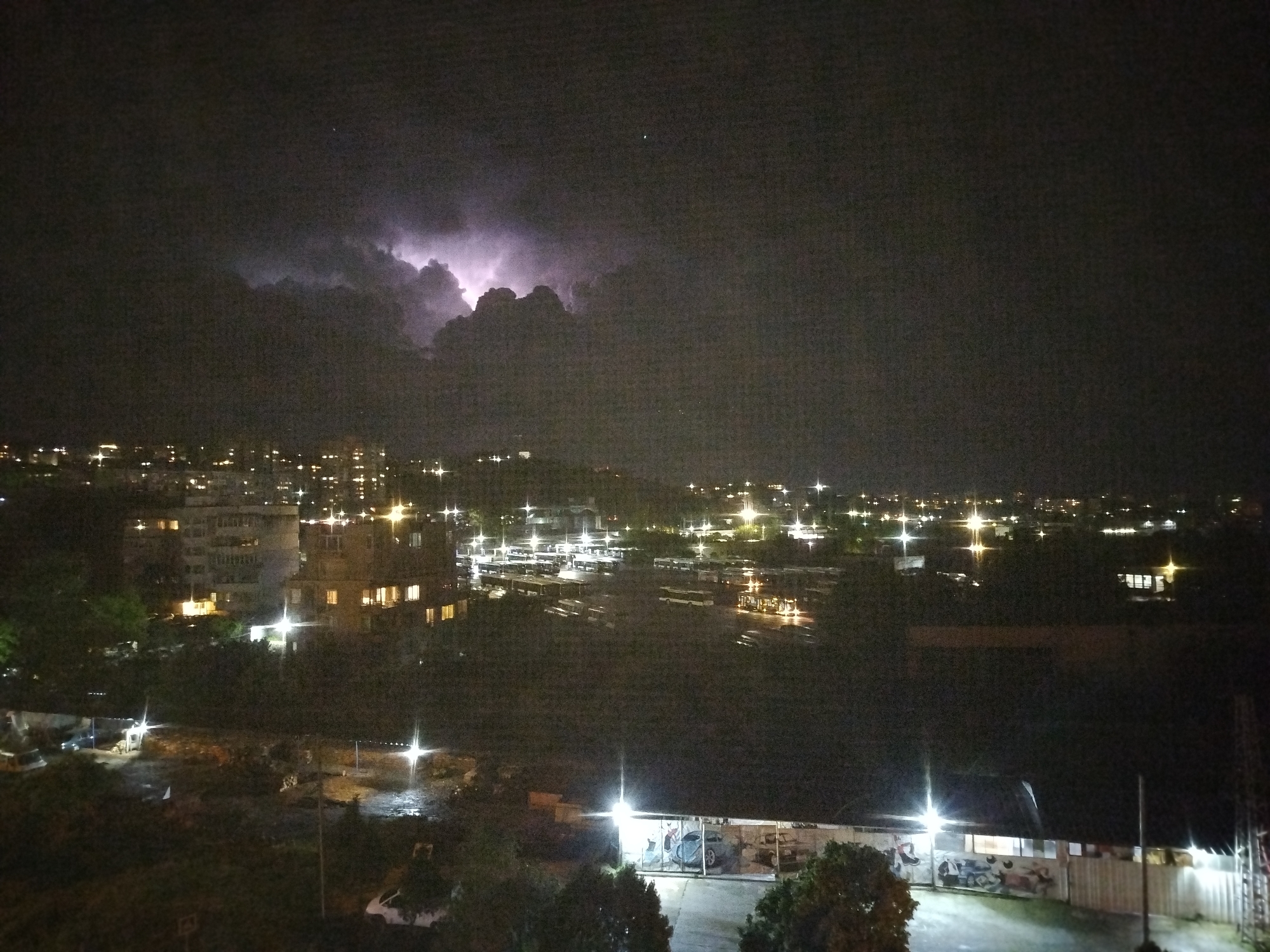 The image shows a nighttime cityscape during a thunderstorm. In the upper part of the image, lightning illuminates dark clouds. Below the clouds, city lights are visible, appearing as scattered bright spots across the horizon. Buildings are interspersed among the lights.
In the middle ground, there are more lights. There also appears to be a parking or storage area for buses.
In the foreground, there's a lower building with bright lights. The image appears to have been taken from an elevated position. Based on the presence of buses, the location might be near a bus depot or transportation hub. The image is taken in Varna, Bulgaria. There are no people visible.