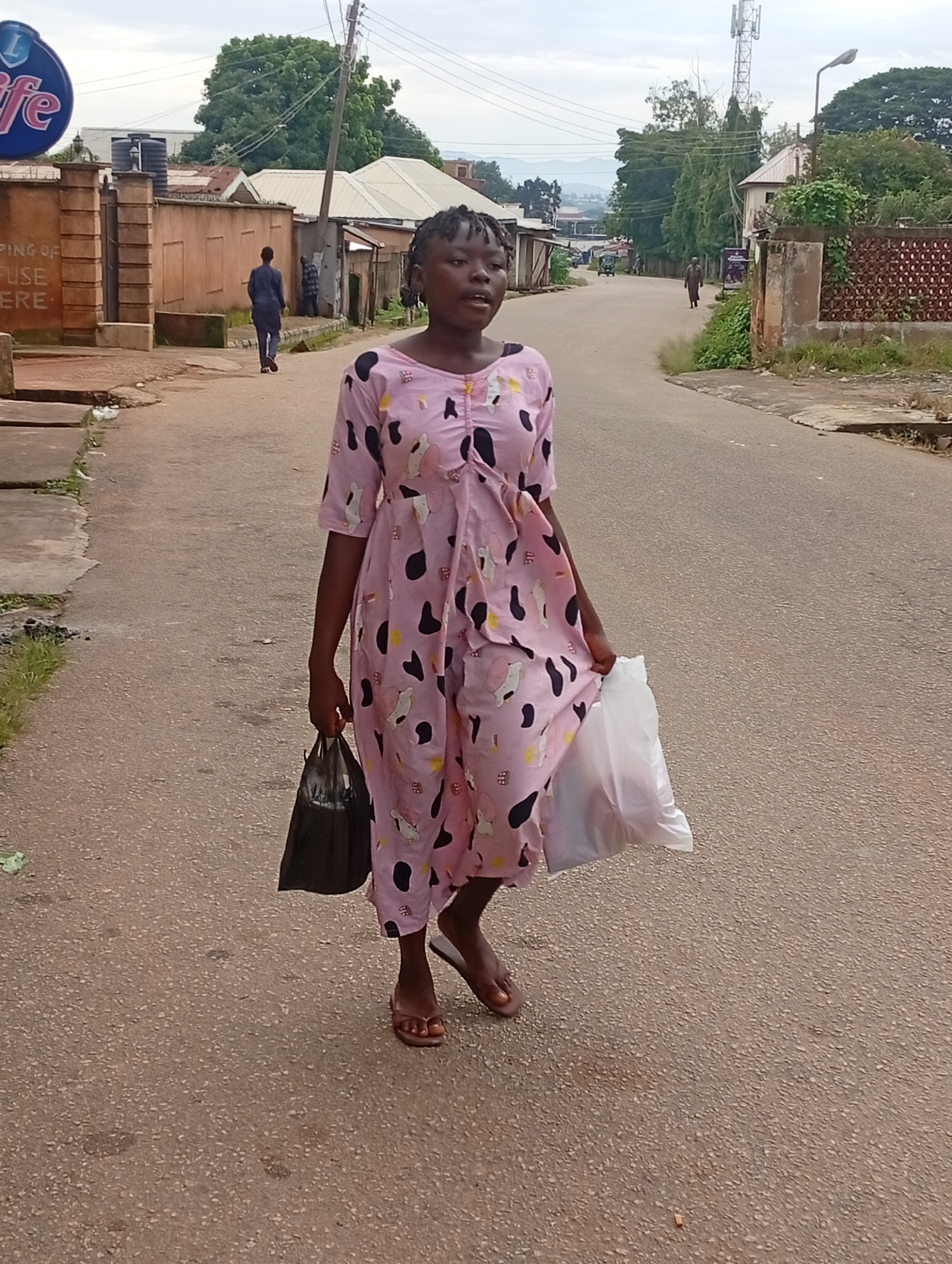 A young Black woman walks down a paved street, holding two plastic bags. She is wearing a pink dress with black and white cartoon-like prints. Her hair is styled in braids. She is wearing sandals on her feet. The street is lined with buildings on both sides, some appearing to be residential, others commercial. Trees are visible in the background. The sky is overcast. In the distance, a cell tower is visible. There is a large blue and white sign with the word "Life" on it to the left of the image. The overall scene depicts an everyday street scene in a populated area.