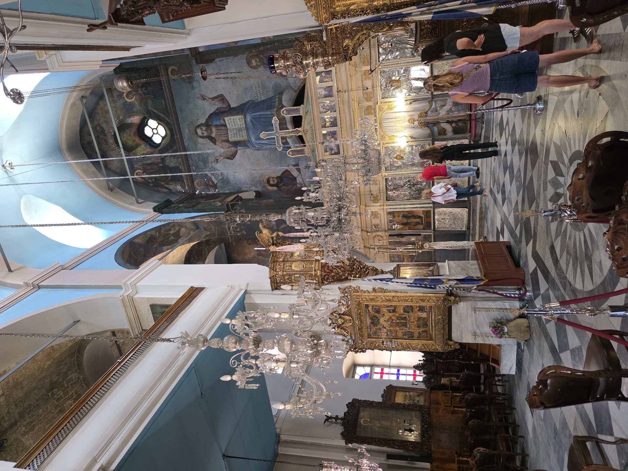 The image shows the interior of a large, ornate church. A blue ceiling with arched structural elements dominates the upper portion of the frame. The walls are adorned with large, colorful murals depicting religious scenes, including a prominent depiction of Jesus Christ. Several large, elaborate crystal chandeliers hang from the ceiling.
In the foreground, the floor is covered in a patterned marble with a diamond motif. Several wooden chairs with carved details are placed around the space. A rope barrier is set up near the center of the image.
In the middle ground, a group of people, appearing to be tourists, are observing the church's interior. They are dressed casually. A large, golden iconostasis, a wall of icons, is a focal point behind them. There are also several large icons in golden frames on the walls.
The background shows more of the church's architecture, including a pulpit and further religious artwork. The overall impression is of a richly decorated and historically significant religious building.
This is the interior of the Church of Agios Titos in Chania, Greece.