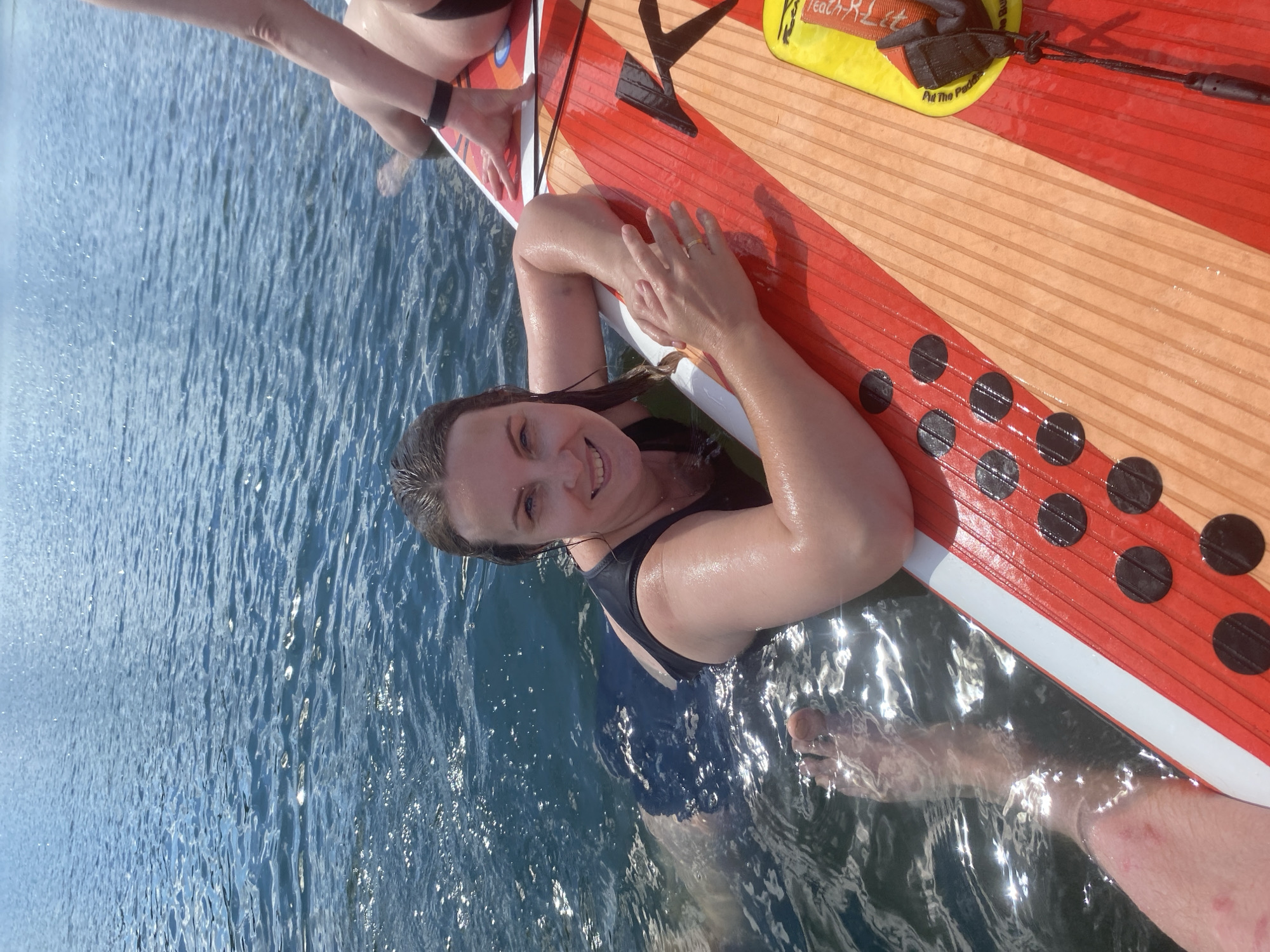 A woman with dark hair, wearing a black swimsuit, is partially submerged in clear blue water, leaning on a paddleboard. Her face is turned towards the camera, and she is smiling. Her arms are resting on the paddleboard, which is red and orange with black polka dots. Another person's legs are visible in the water to the left of the woman. The water is calm with some ripples and sunlight glinting on its surface. The paddleboard has a yellow sticker with text that is partially visible. The environment appears to be a natural body of water, possibly a lake or river. The image does not contain any identifiable landmarks or clues to suggest it is located in Novyi, Russia.