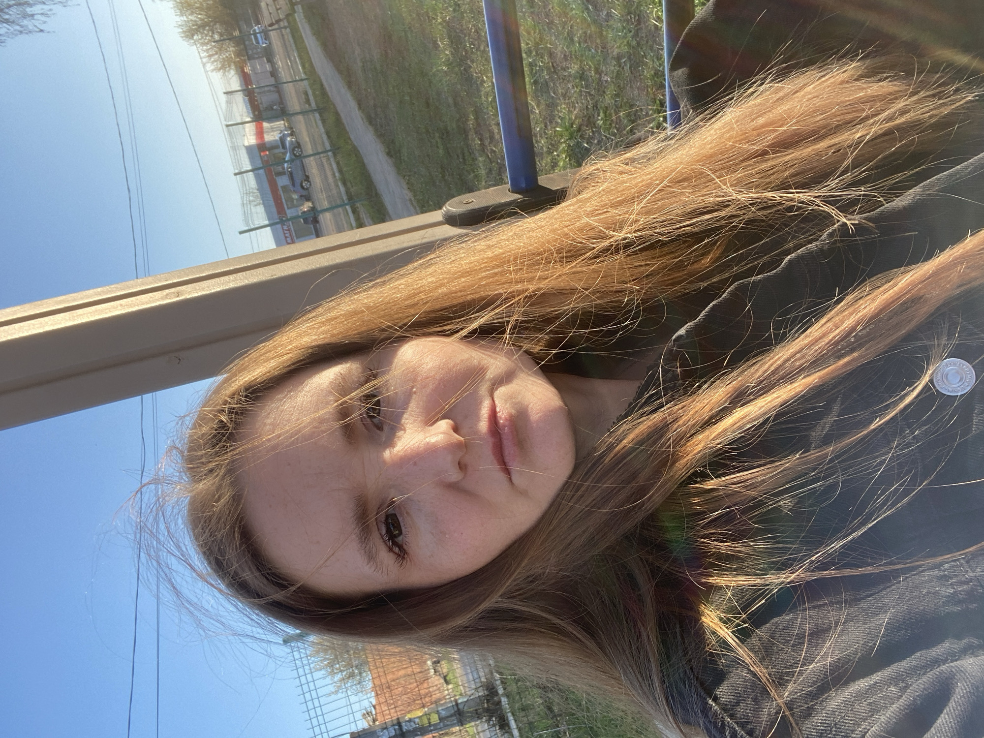 A young woman is seated at what appears to be a bus stop in Chaikovsky, Russia, as suggested by the surrounding environment. She is wearing a dark-colored jacket. The background includes greenery and a building enclosed by a fence. The sky is clear and blue. Electrical lines are visible in the sky.