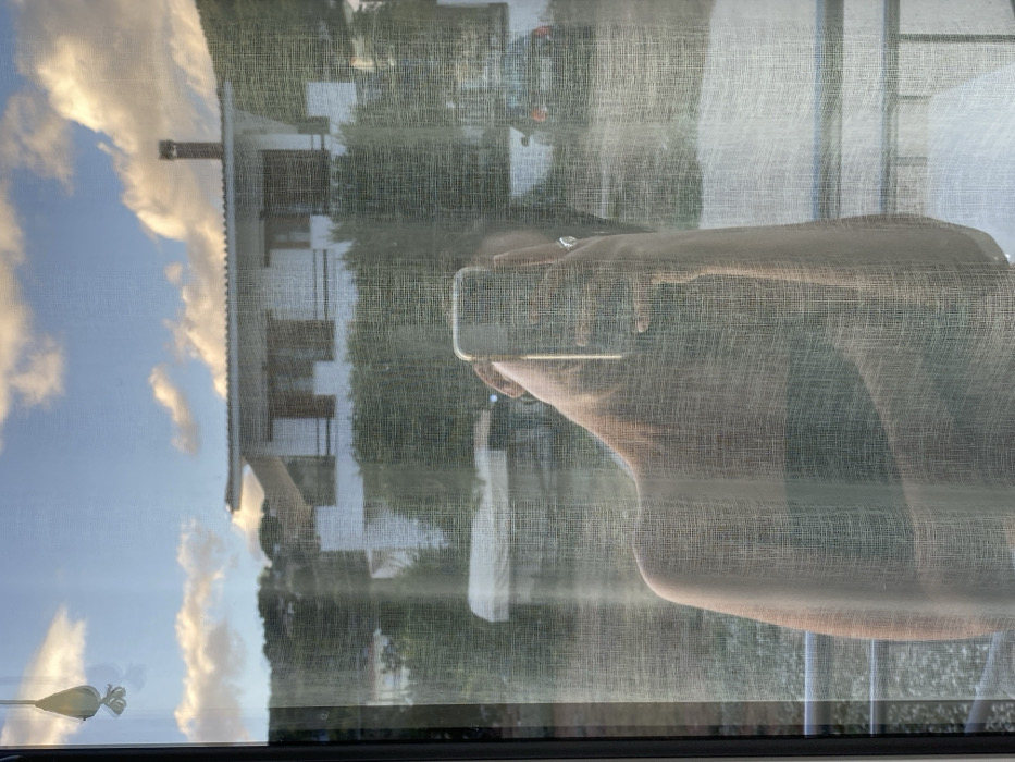 The image displays a reflection self-portrait captured through a translucent, textured curtain from an elevated vantage point in Skala, Greece. A person's upper torso and right arm are visible in reflection, holding a silver smartphone vertically to photograph the scene; a ring is present on the right hand. The external environment, visible beyond the curtain, depicts a daytime setting under a blue sky with scattered white cumulus clouds.
In the mid-ground, a white two-story building with a distinctive curved tile roof and a dark chimney is prominent. The building features multiple windows, some with dark shutters or frames, and appears to have balconies. Lush green vegetation, including trees and shrubs, surrounds the building and extends up a hillside in the background. In the lower background, a light-colored, covered boat is partially visible to the left, and two vehicles are parked: a lighter-colored van or car to the left and a darker sedan to the right. A metal balcony railing is discernible in the lower right foreground. A small, pale, teardrop-shaped decorative item hangs suspended in the upper left corner of the frame, partially visible through the curtain.