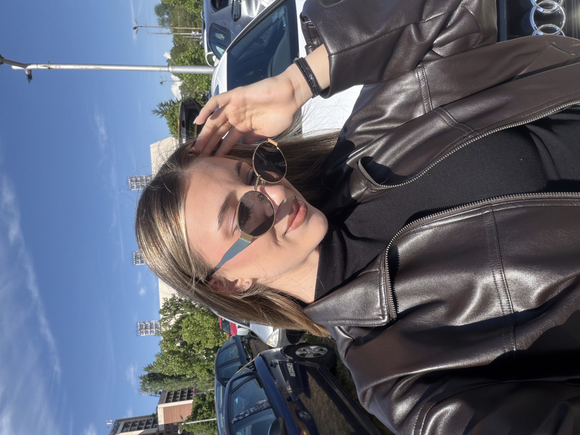 A young woman poses in an outdoor parking lot. She is wearing a brown leather jacket over a black top, and sunglasses with a rounded shape. Her blonde hair is down. She is wearing a dark-colored bracelet on her left wrist. Behind her, various cars are parked. In the distance, there are buildings and trees under a blue sky with some clouds. Based on the style of architecture and street features visible, the location is identified as Sofia, Bulgaria.
