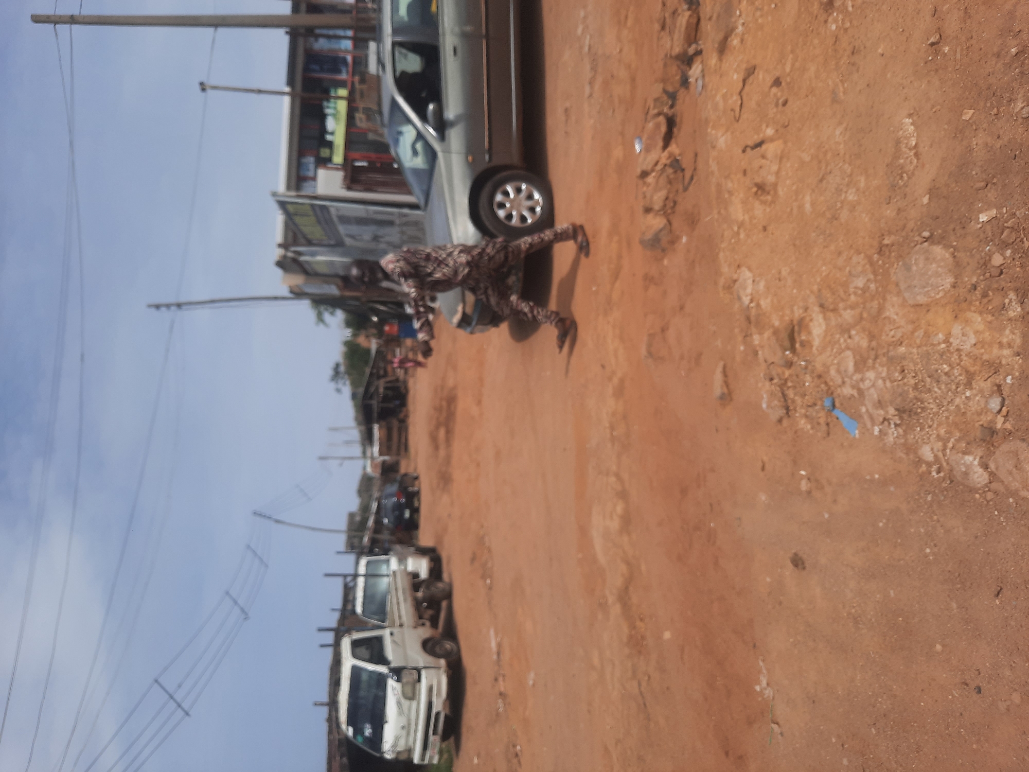 A person wearing a brown and black patterned outfit is squatting near the front tire of a silver car on a dirt road in Gura Top, Nigeria. Further down the road are several white vans. There are also buildings and power lines visible in the background.