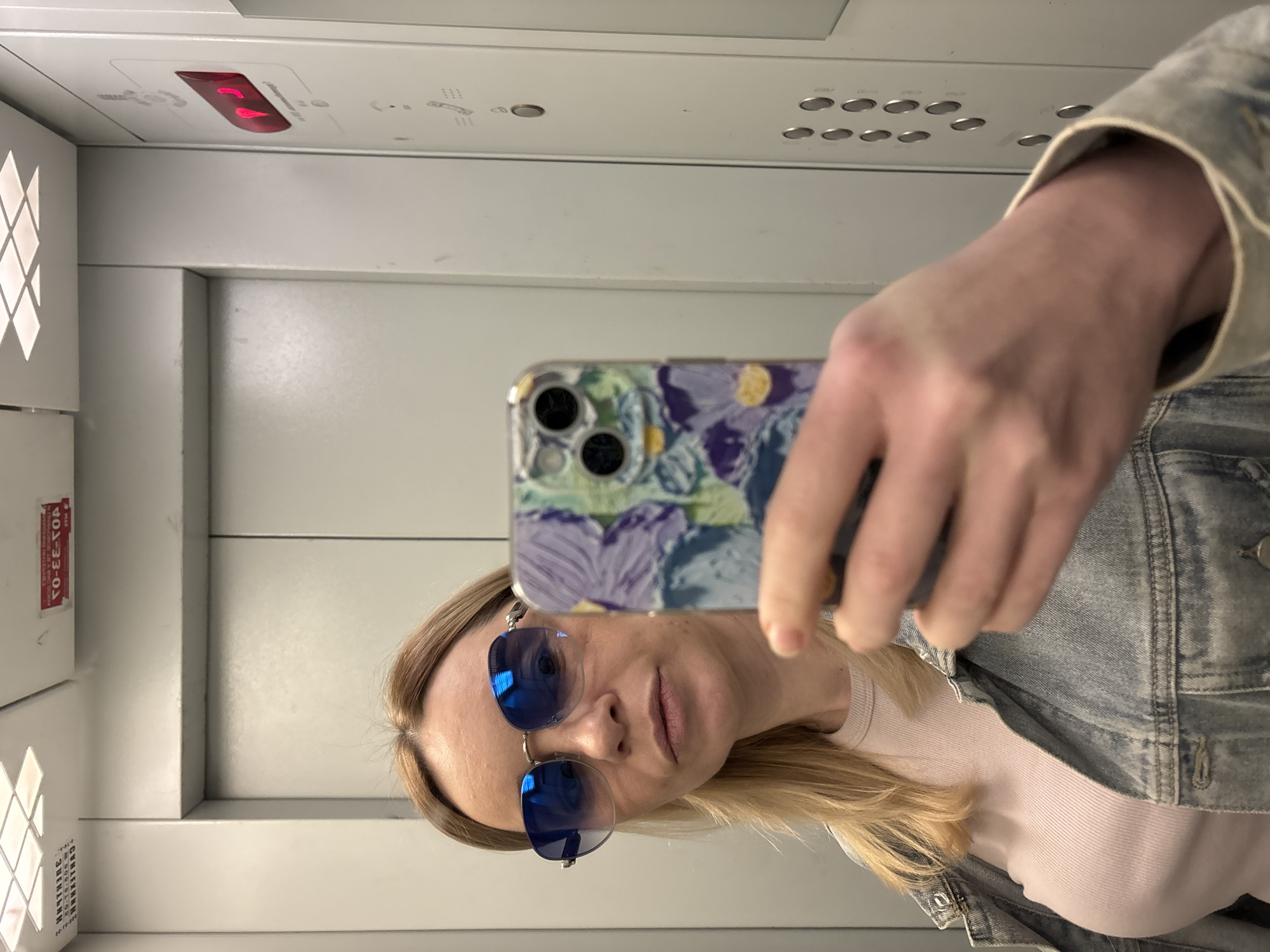The image, originally rotated 90 degrees clockwise, depicts a mirror selfie taken by a woman in an elevator. The woman, with blonde hair, is seen from her forehead to her chest, wearing blue mirrored sunglasses, a light-colored ribbed top, and a light blue denim jacket. She holds a smartphone with a distinctive purple, green, and yellow patterned case to capture the reflection.
The elevator interior features light grey or white panels. To the upper left, a white panel with a diamond-patterned cutout is visible, likely for ventilation or lighting. A red and white rectangular warning label with Cyrillic text is affixed vertically to the left wall. Visible text on the label includes "ПРЕДУПРЕЖДЕНИЕ!" (WARNING!), "СТОП", "401-33-02", and "ЗАПРЕЩАЕТСЯ" (FORBIDDEN).
Above the woman's head and to her right, an elevator control panel features multiple round silver buttons and a prominent red indicator light. The red light displays two glowing, indistinct red segments, possibly indicating a floor or status. The overall setting appears utilitarian. The Cyrillic text on the warning label strongly suggests the location is in Russia. The woman's expression is neutral, partially obscured by her sunglasses.