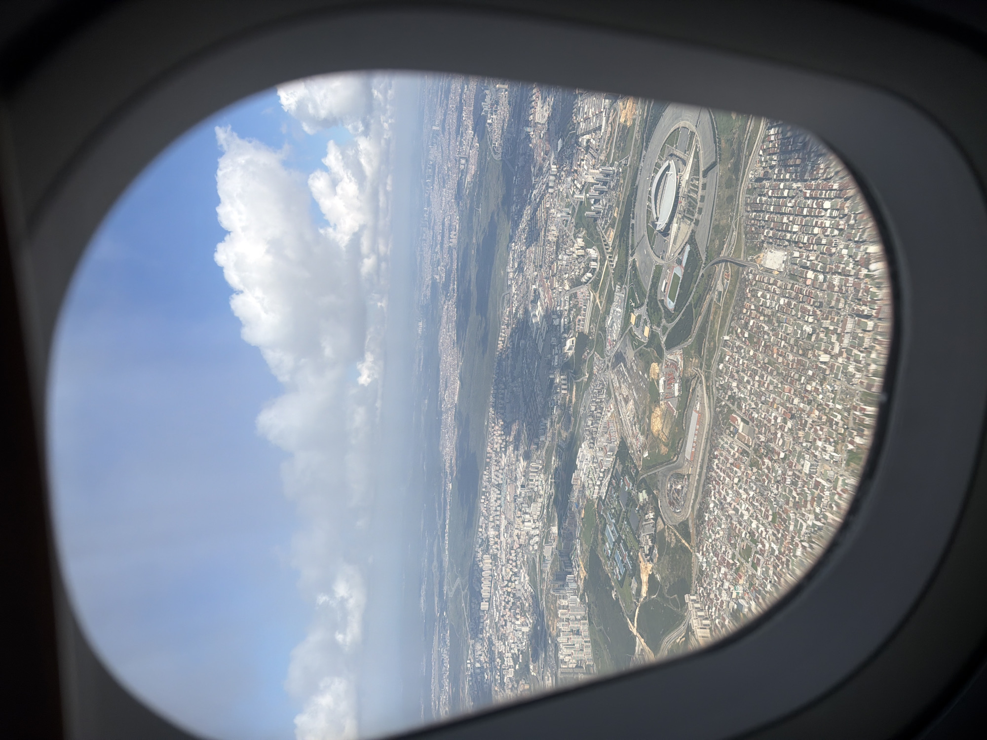 The image presents an aerial view of an expansive urban landscape as seen through an airplane window. The dark gray, oval window frame partially obscures the top, bottom, and left edges of the scene. The perspective is rotated clockwise, showing the sky on the left and the ground on the right.
The sky is a clear blue, adorned with large, fluffy white cumulus clouds that are brightly lit, indicating daytime. Below, the ground is dominated by a vast cityscape stretching into the distance. A prominent feature is a large, circular stadium complex located towards the upper right of the frame. This stadium has a distinctive white, possibly domed or retractable, roof and is surrounded by smaller sports fields, paved areas, and a network of access roads.
Beyond the stadium, the city sprawls with countless buildings, mostly appearing residential with varying roof colors, interspersed with larger commercial or industrial structures. An intricate web of roads and highways is visible, connecting different parts of the urban fabric. There are also patches of darker green vegetation, suggesting parks or undeveloped areas within the dense concrete environment. The weather appears clear with good visibility, under bright sunlight, suggesting a late morning or early afternoon time. No discernible text is visible within the image.