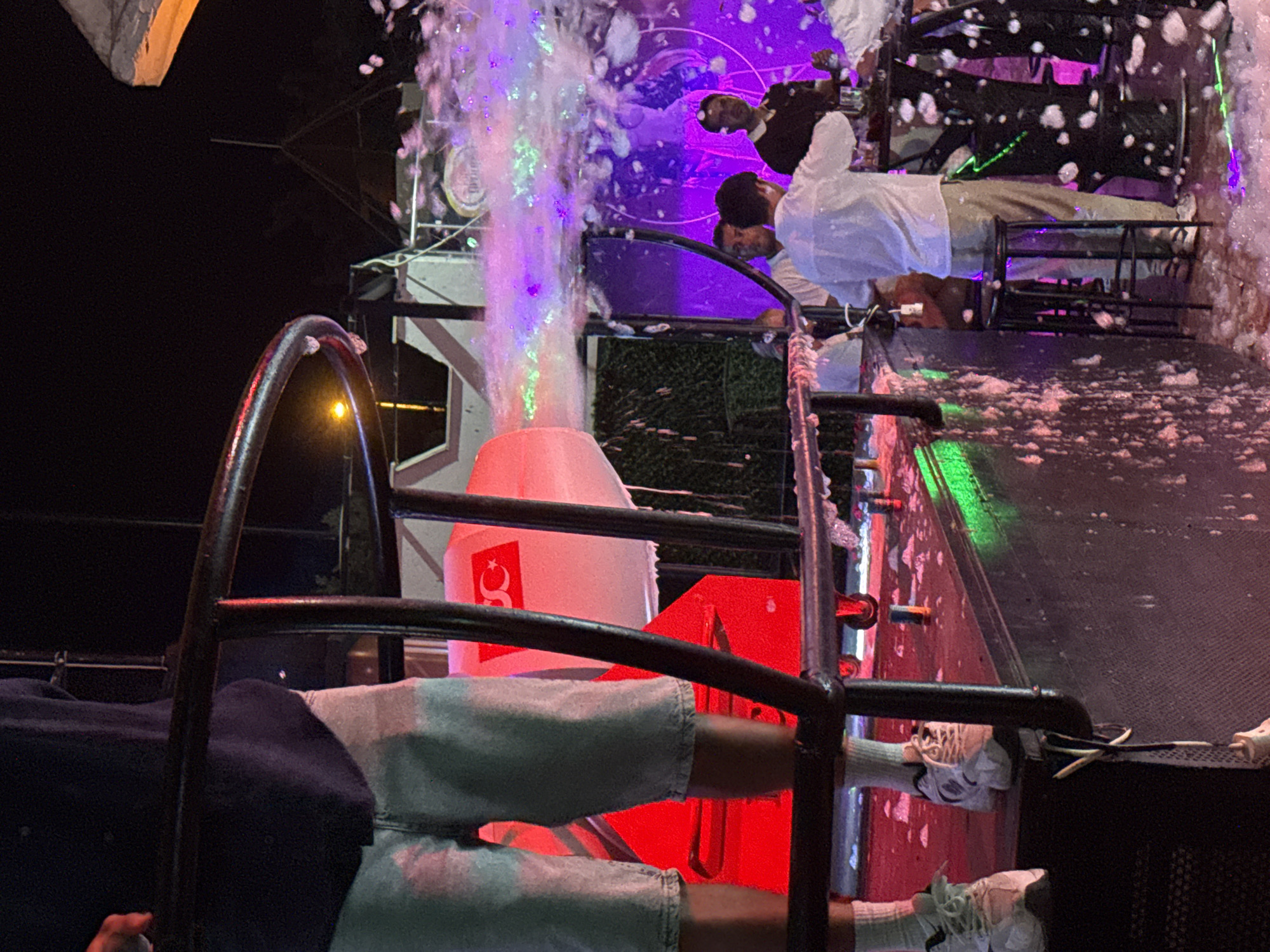 The image depicts a nighttime scene at a beach club or bar in Alanya, Turkey, likely during a foam party. In the foreground, a person is standing on a raised platform, wearing light-colored shorts, white socks, and athletic shoes. To their right, a large, industrial foam cannon is visible, emitting a dense cloud of white foam illuminated by purple and green lights. The foam is spreading across the area, covering the ground and some surfaces.
In the background, several individuals are present. One person in a white shirt is standing behind a bar or counter, appearing to be serving drinks or interacting with patrons. Other people are also visible, some standing and others possibly seated on stools. The environment is lit by various colored lights, creating a festive and energetic atmosphere. A large red object with a Turkish flag symbol is partially visible on the left side of the frame, suggesting it may be part of the club's decoration or equipment. The overall scene conveys a lively nightlife experience with music, dancing, and the characteristic foam from the party.