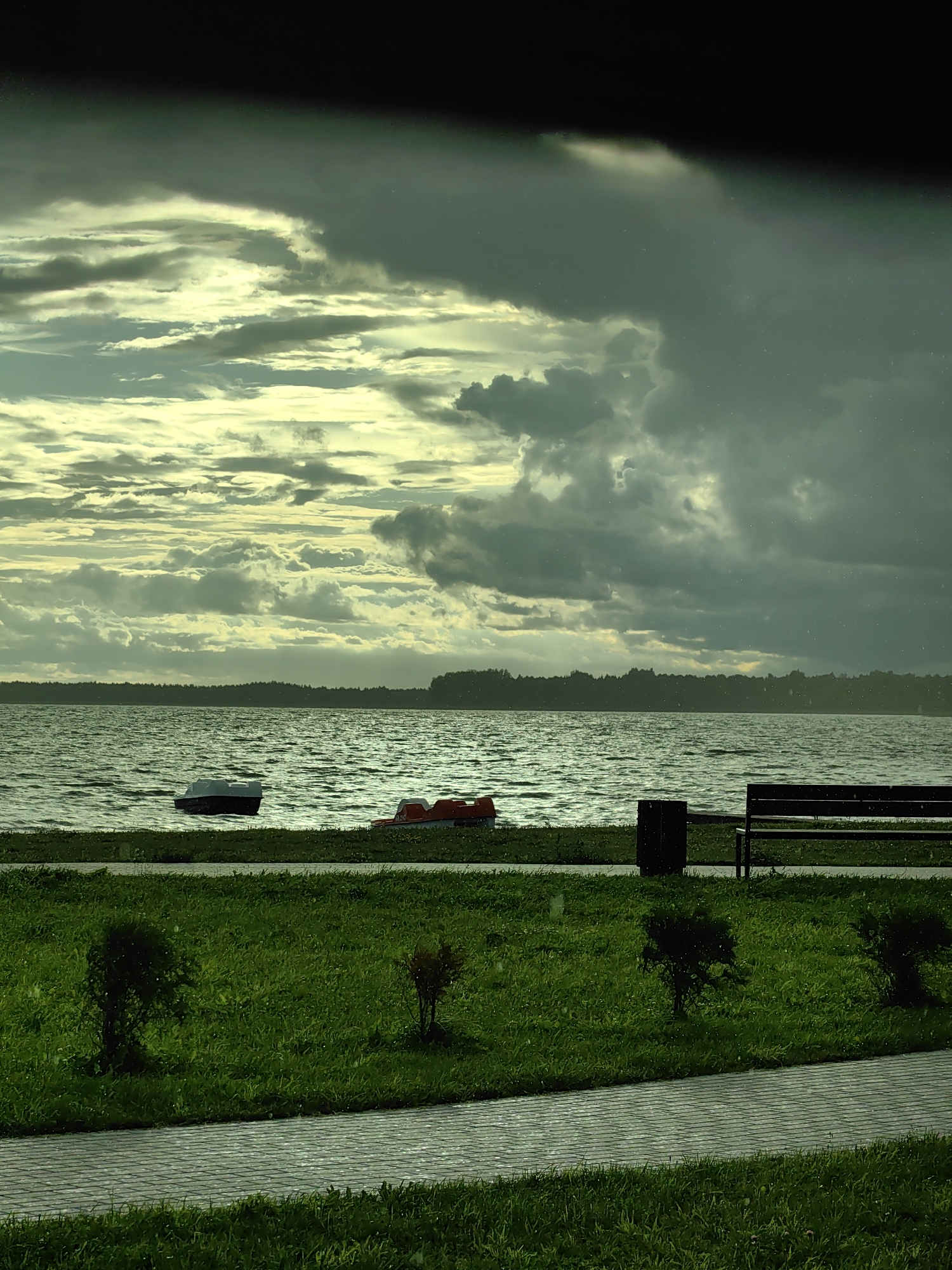 The image depicts a scene by a lake in Myadzyel, Belarus. The sky is dramatic, with a mixture of dark, heavy clouds and lighter, illuminated areas, suggesting an overcast or stormy day. The lake's surface is rippled, indicating a gentle breeze or waves. In the water, two pedal boats are visible near the shore: one white and dark, and another orange and red. The shoreline features a strip of green grass, a paved walkway, and a park bench with a trash bin. Small bushes are planted on the grassy area. The overall atmosphere is serene yet moody, possibly due to the weather.