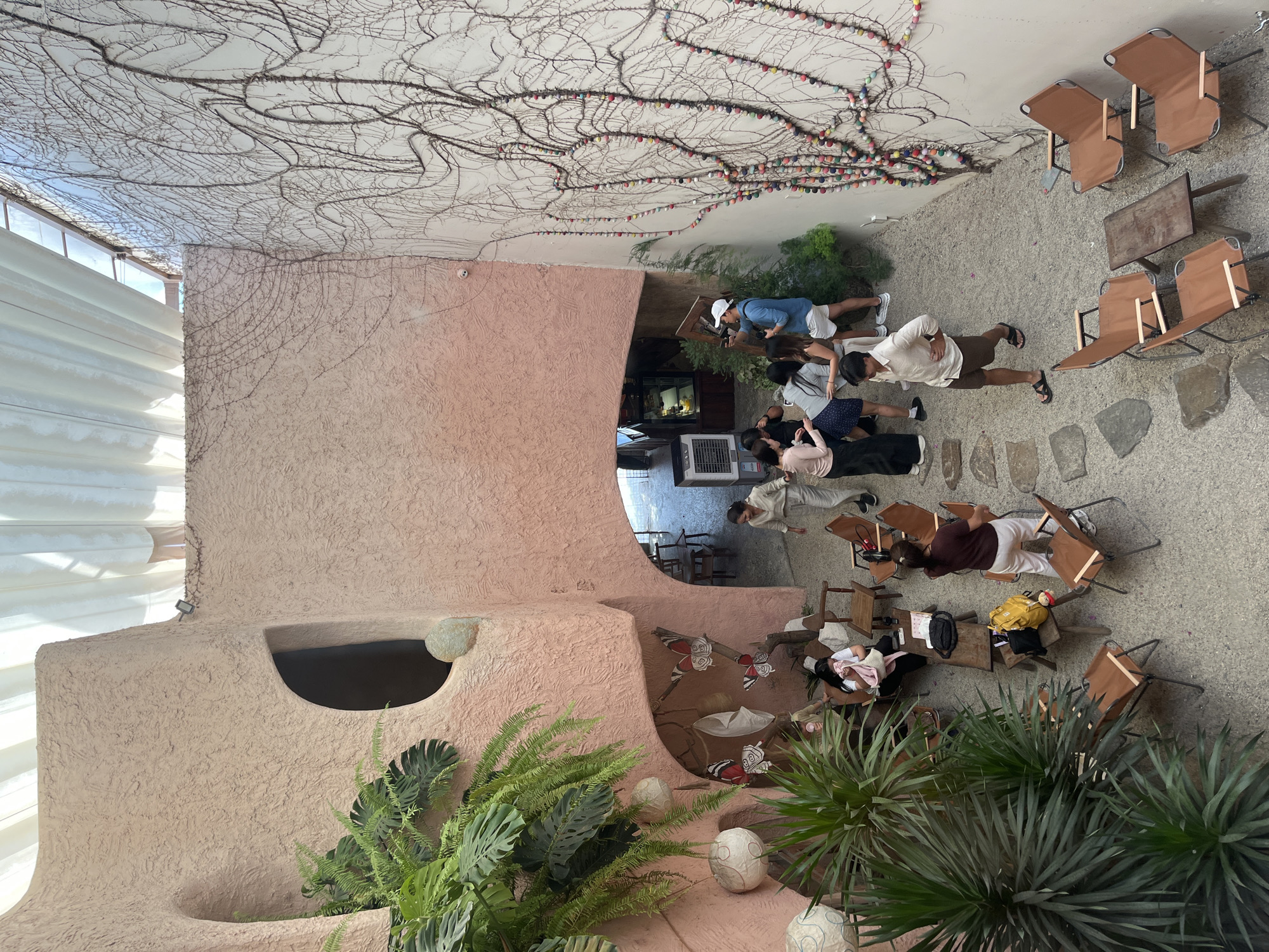 The image captures a lively outdoor courtyard of a cafe in Nha Trang, Vietnam, characterized by its unique organic architecture and lush greenery. The walls are a textured, pale pink, with rounded archways and openings that give the space a cave-like feel. Large potted plants, including ferns and palm-like foliage, are strategically placed throughout the courtyard, softening the architectural lines. Decorative paper lanterns hang from branches, and artificial butterflies adorn a niche in the wall.
The ground is covered with small, light-colored pebbles, interspersed with larger, flat stones forming informal pathways. The courtyard is filled with various seating arrangements, including individual portable chairs and small wooden tables, suggesting a relaxed atmosphere conducive to socializing or enjoying refreshments.
A group of people are scattered throughout the space. In the foreground, a woman in a dark top and white pants stands near some chairs. To her left, another woman is seated, holding a pink cloth. Further back, near an arched entryway, a cluster of individuals is engaged in conversation. One man in a blue shirt and white shorts holds a phone, possibly taking a picture. Another man in a light-colored shirt and brown shorts walks across the courtyard.
The overall impression is that of a trendy and aesthetically pleasing cafe that blends natural elements with distinctive architecture, creating an inviting urban oasis. The scene suggests a typical day at the cafe, with patrons enjoying the ambiance and each other's company.