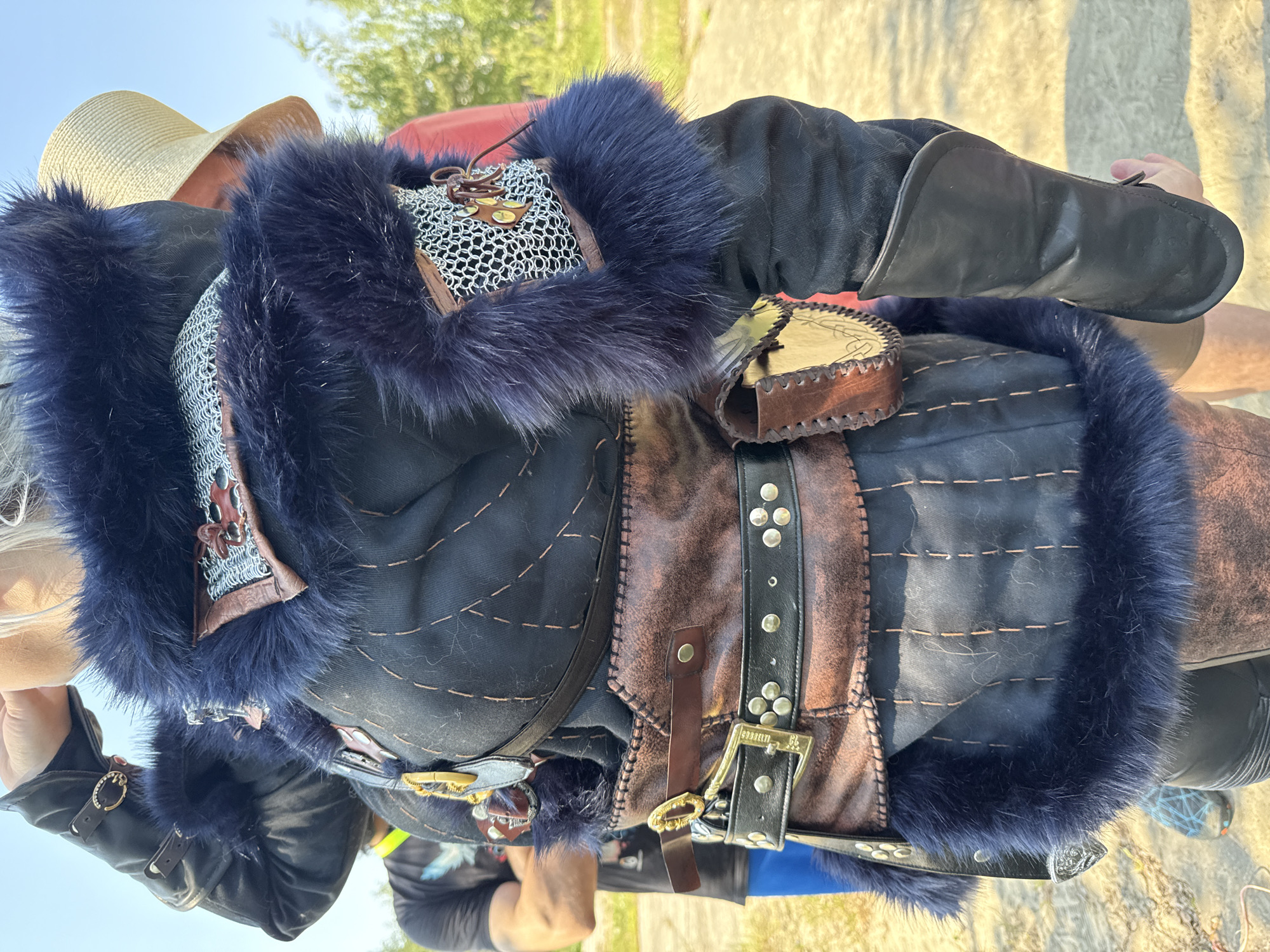 A person is wearing a heavily detailed outfit consisting of a dark jacket trimmed with blue fur, over which is worn a brown leather belt adorned with silver studs and a large buckle. Protruding from the jacket's shoulder is a section of chainmail. The location is possibly Kotelniki, Russia. The environmental details suggest an outdoor setting. The person is wearing a beige hat.