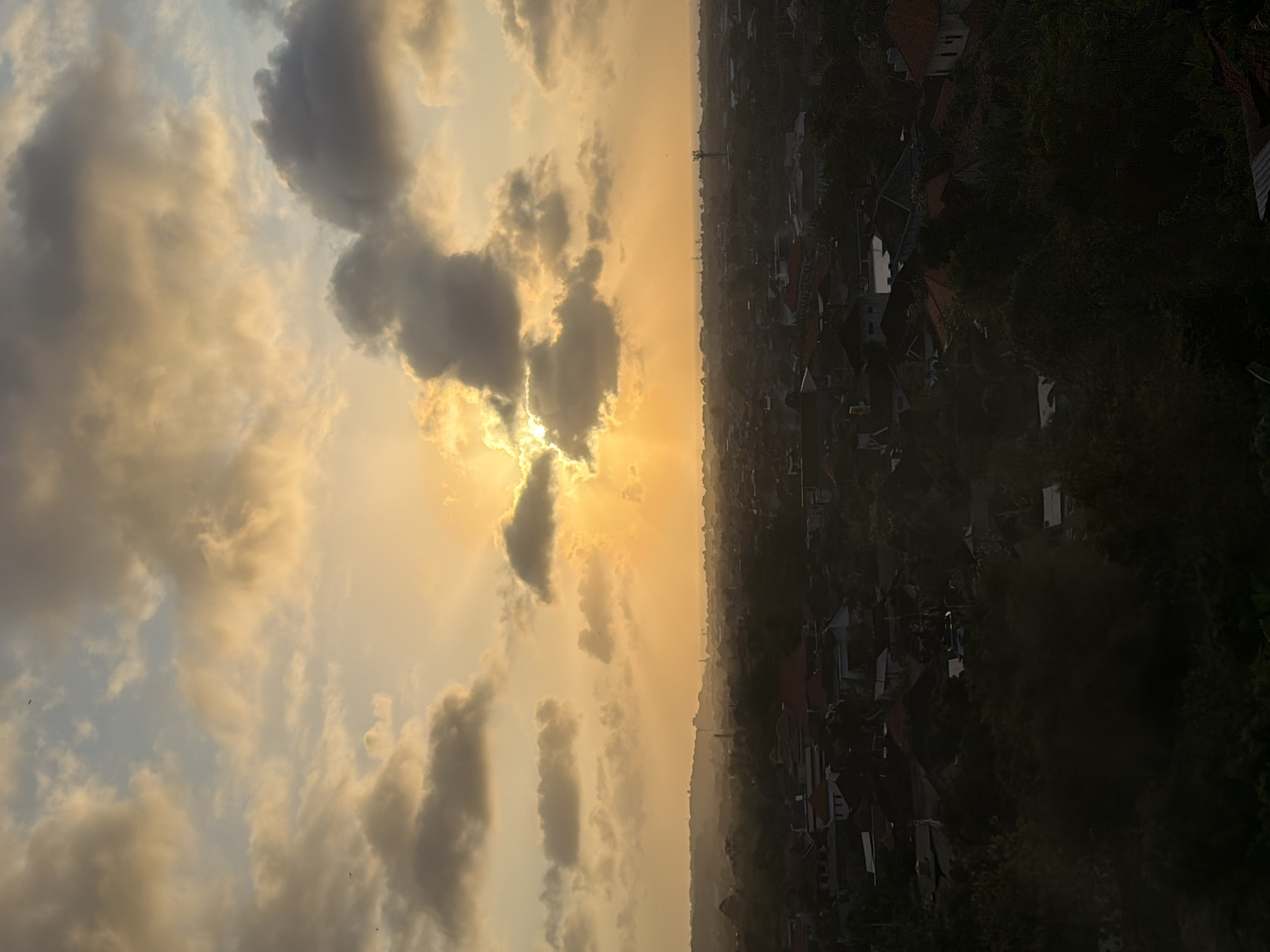 The image depicts a sunset over the city of Kuta Selatan, Indonesia. The sky is filled with dramatic clouds, some dark and heavy, others illuminated by the golden rays of the setting sun, creating a light show of sunbeams piercing through the atmospheric haze. Below the sky, the city sprawls out, a dense collection of buildings with red-tiled roofs nestled amongst lush, dark green foliage. The overall impression is one of a peaceful, residential area bathed in the warm, fading light of day, with the distant horizon blurred by the atmospheric conditions.