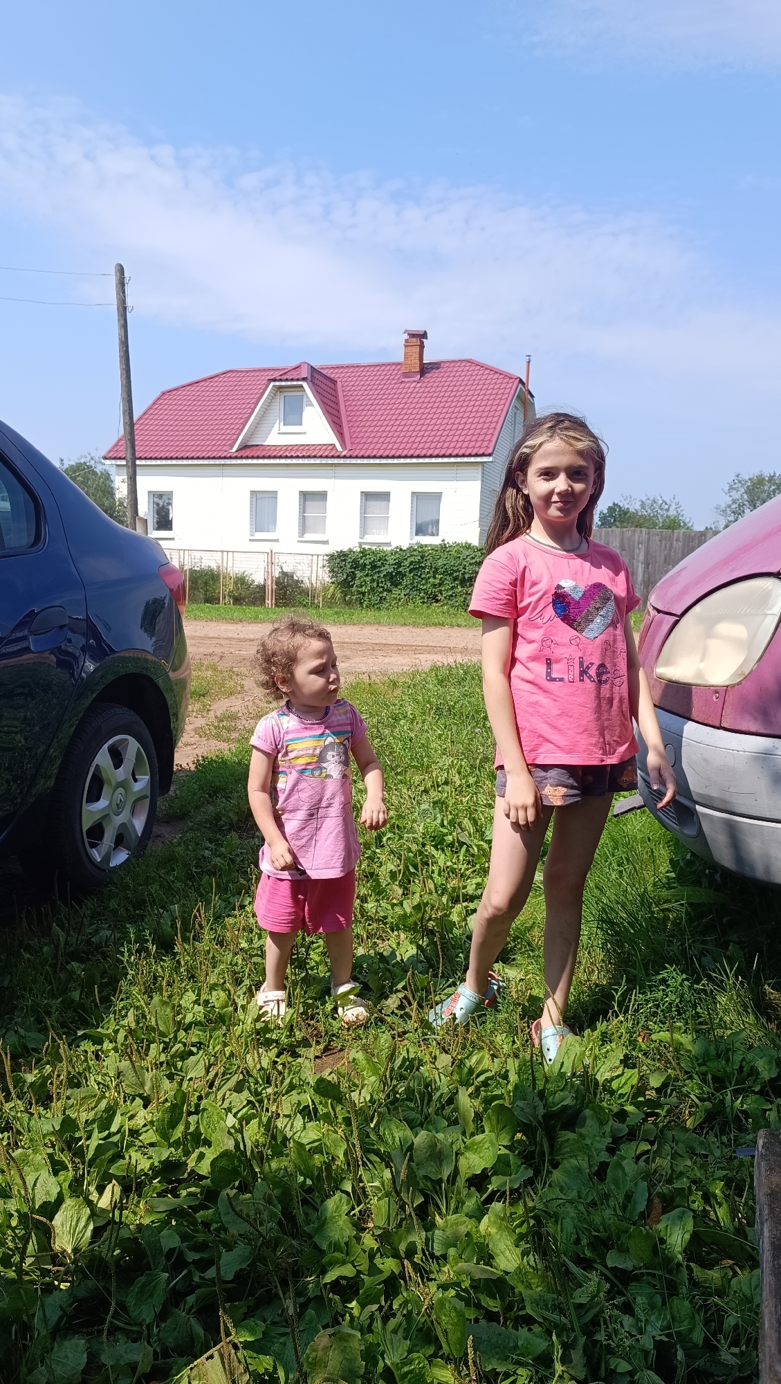 Two young girls are standing in a grassy area with a white house with a red roof in the background. The girl on the left is shorter with curly brown hair and is wearing a pink t-shirt and pink shorts. The girl on the right is taller with long brown hair and is wearing a pink t-shirt with a heart design and dark shorts. To the left of the girls is the rear of a dark blue car. To the right of the girls is the front of a pink car. The house appears to be a residential building. The overall environment suggests a rural or suburban setting, possibly in Karsha, Russia, given the context. The sky is clear and blue with a few wispy clouds. The lighting indicates it is daytime.