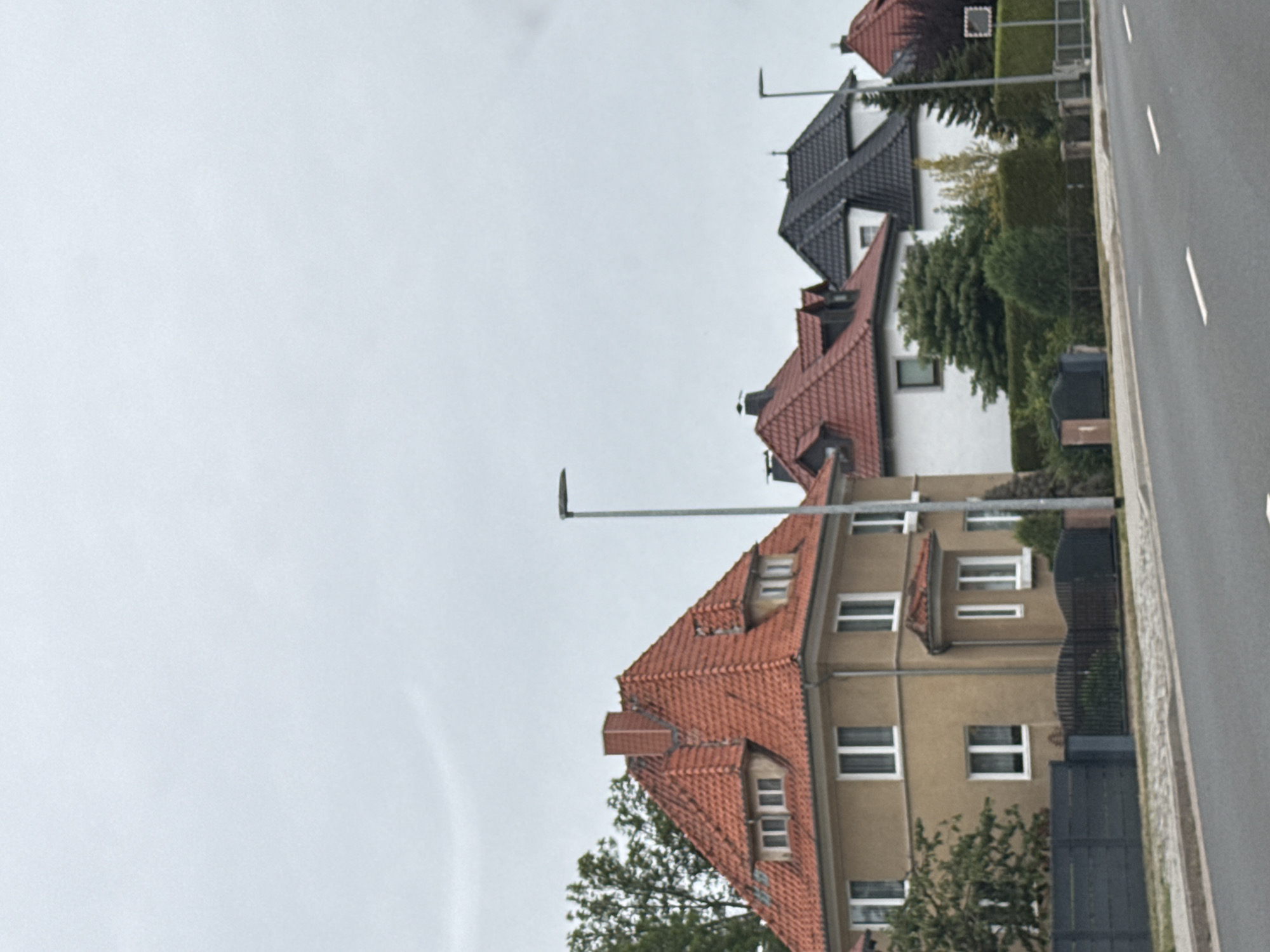 The image depicts a street scene in Nordhausen, Germany, featuring a row of residential houses along a paved road. The sky is overcast. The houses vary in architectural style and color, with red tiled roofs being a prominent feature on several of them.
On the left side of the image, a light brown house with a steeply pitched red tiled roof and dormer windows is visible. It has white-framed windows on the upper floors. A dark gray fence runs along the front of this property. Adjacent to this house, to its right, is a white house with a red tiled roof, also featuring dormer windows. Further to the right, a house with a black tiled roof and white walls is visible. Additional houses with red tiled roofs are seen further down the street, suggesting a residential neighborhood.
Several streetlights are present, one tall gray pole with a modern lamp is prominently located to the right of the first house. Other street lamps with simpler designs are also visible along the street. Trees and green hedges line the properties. The road in the foreground has white lane markings. There are no people or vehicles visible in the image. The overall impression is a quiet, suburban street on a cloudy day.