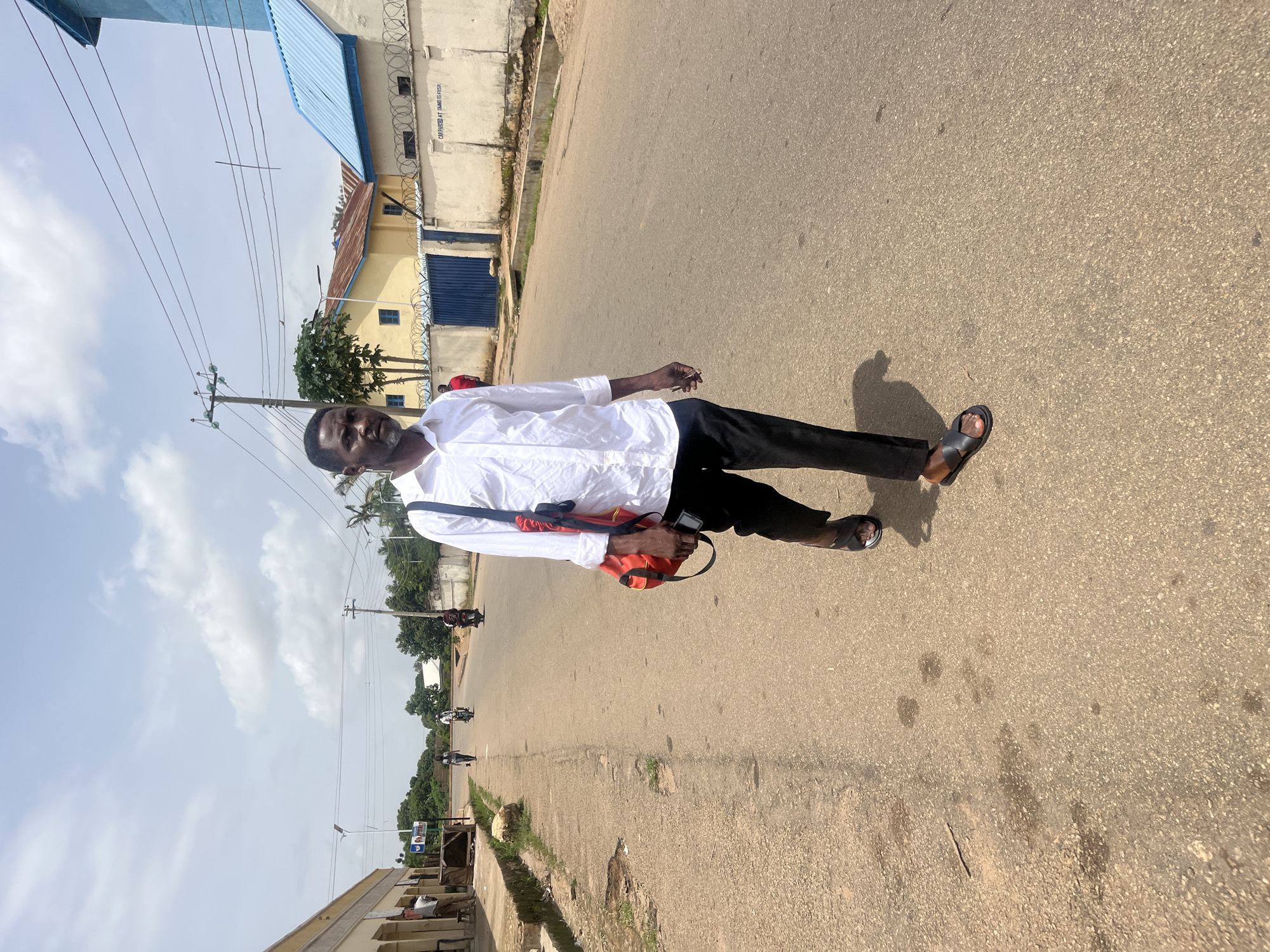 A man with dark skin, a beard, and a receding hairline is walking towards the camera on a paved road in Bukuru, Nigeria. He is wearing a white long-sleeved collared shirt, black trousers, and dark sandals. He has a red bag with a black strap slung over his right shoulder. The road is asphalt and appears well-maintained, with some oil stains and water marks. On the left side of the road, there is a sidewalk with some vegetation and buildings. On the right side of the road, there is a wall with barbed wire and a blue gate, with buildings behind it. The sky is partly cloudy. In the background, there are a few other people and motorcycles. The lighting suggests it is daytime.