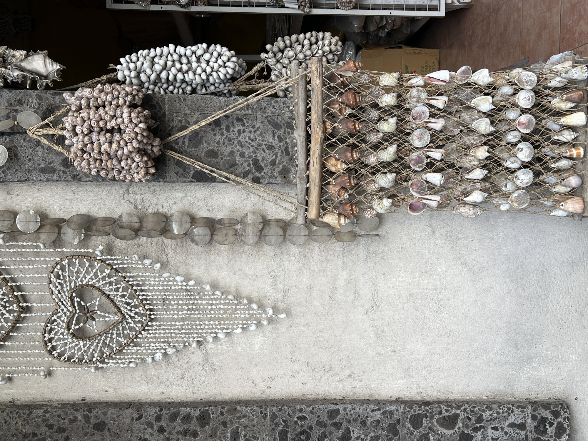 The image displays a vibrant collection of seashell crafts, likely presented as merchandise in a shop or market stall within the Ubud District, Indonesia. The scene is viewed from an slightly elevated perspective, showcasing the intricate details of the handcrafted items.
In the upper section, two large, elongated clusters of small shells are prominent. One cluster consists of pale, greyish-white shells, while the other features smaller, spiraled shells in natural beige and brown tones, both bundled together with coarse twine. Adjacent to these, a rustic display made of wooden sticks and twine netting holds a variety of larger seashells, including conch and bivalve shells, showcasing their diverse shapes and natural colors from white to brown and purple.
Below these displays, a horizontal garland of flat, translucent circular shell pieces is visible. Further down and to the left, two decorative heart shapes are crafted from woven material, possibly rope, and intricately adorned with numerous small white shells. The visible heart features a small, star-shaped shell at its center. Strands of tiny white shells extend from these hearts, forming a flowing, triangular pattern.
The background is generally dark, suggesting an indoor or shaded environment, with glimpses of metal shelving and a partially visible cardboard box. Text on the box reads "TATI NAAMIH NAAT NUUTH". The entire display rests against a textured, light grey wall or concrete surface. No people are visible, and no specific activity is taking place beyond the static presentation of these handcrafted items.