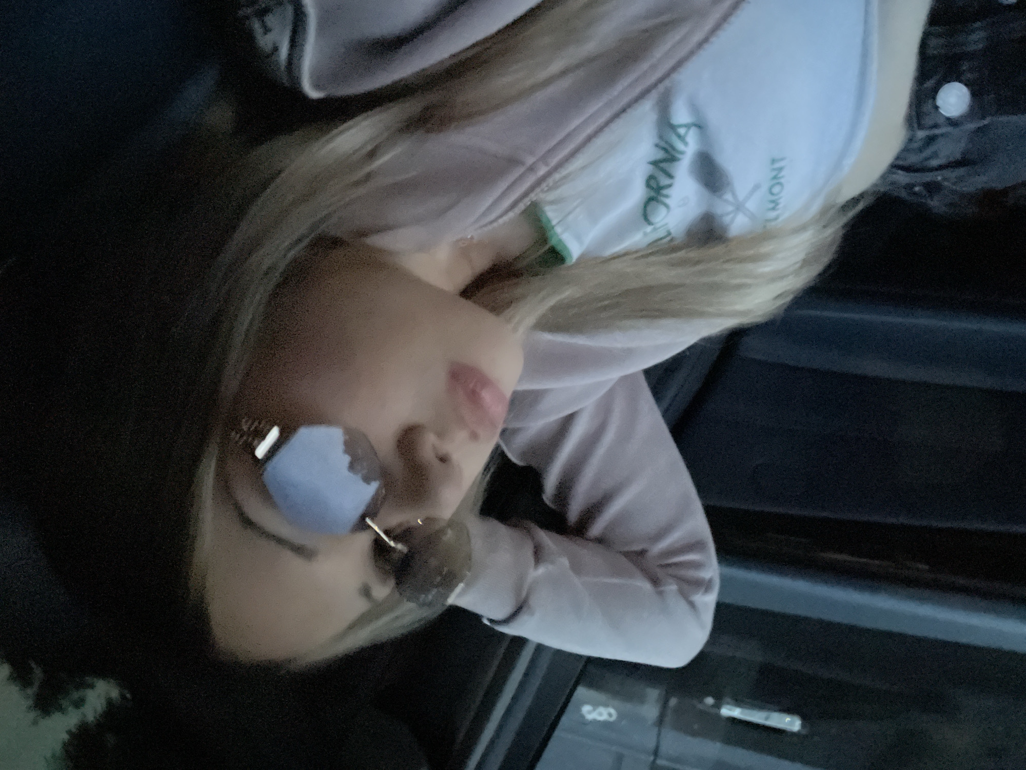 A young female, characterized by blonde hair and pink-tinted lips, is positioned within the interior of a vehicle, in a reclined posture with her right arm bent behind her head. She is wearing rimless sunglasses with reflective, light-blue lenses that reflect ambient light and indistinct dark shapes consistent with external foliage or structures. Her attire consists of a light-colored tank top with green lettering, partially identifiable as "LOMONT" and "CALIFORNIA," beneath an open, light pink or lavender zip-up hoodie. A dark-colored lower garment, possibly jeans, with a visible button, is partially visible. The vehicle's interior is dim, showcasing a dark door panel on the left with a horizontal trim and a metallic door handle. The image contains no visual evidence, such as landmarks, signage, or specific vehicle identifiers, to confirm the location as Dobrich, Bulgaria.