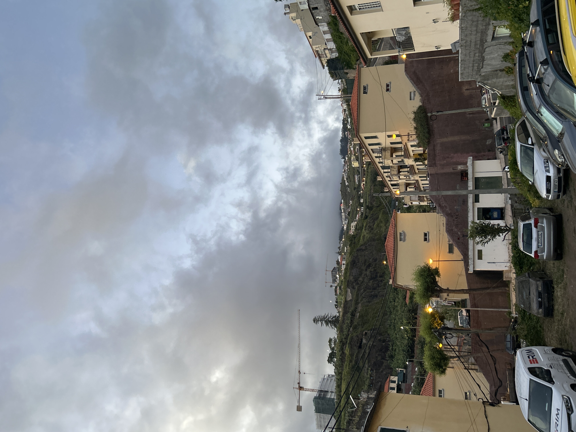 The image depicts a cityscape on a hillside under an overcast sky. It is likely that the location is Câmara de Lobos, Portugal. In the foreground, there's a street with parked cars, including a white van with "ERIM" printed on its side. Houses and apartments, primarily beige with red tile roofs, are built into the hillside. Streetlights are visible amongst the trees and buildings. In the distance, there are construction cranes, hinting at ongoing development in the area. The sky is covered with grey clouds, suggesting a rainy or cloudy day. The focus is on the built environment and the natural landscape.