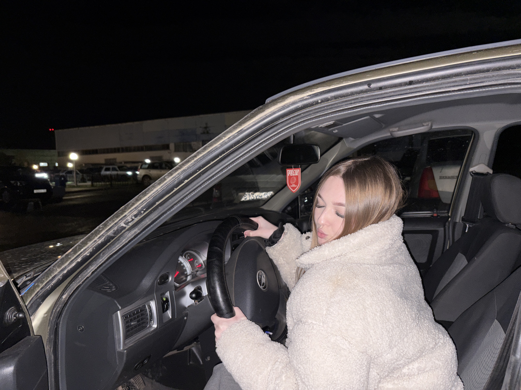 A young woman is seated in the driver's seat of a car at night, viewed from the open driver's side door. She has light brown hair and appears to have her eyes closed or cast downwards. She is wearing a thick, cream-colored, textured jacket (possibly fleece or sherpa) and is holding the steering wheel with both hands, which has a black cover on it.
The car's interior features a dark dashboard with an instrument cluster showing red needles and white markings, and air vents with silver trim. A red air freshener with the word "FELIX" is visible hanging from the rearview mirror.
Outside the car, the scene is dark, indicating nighttime. Several streetlights illuminate a parking area with other vehicles, including a car with "RENAULT" visible on its rear. A large, light-colored building with windows is present in the background. Part of a Russian license plate, "M893XM 159", is visible on a vehicle in the distance. A yellow/orange traffic cone can also be seen through the rear window. The overall setting appears to be an urban or industrial area at night, likely a parking lot, in Chaikovsky, Russia. The woman's closed eyes and posture suggest a moment of rest, contemplation, or posing.