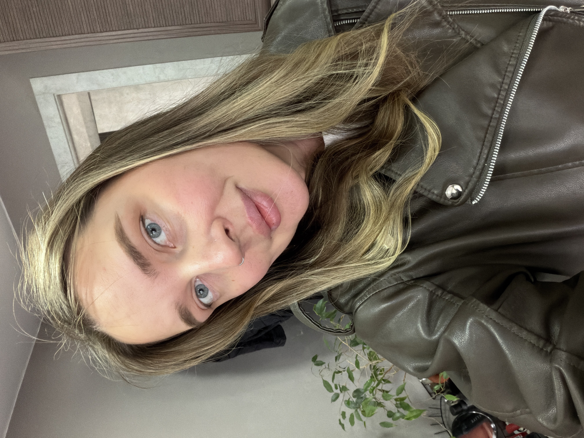 A fair-skinned woman with light blue eyes and blonde hair, wearing a brown leather jacket, is captured in a medium-shot portrait. Her hair is styled with loose waves. She appears to be indoors. A small plant with green leaves is visible near the bottom of the frame. A light gray wall and a door frame are partially visible in the background. This does not provide any information to determine it is in Chaikovsky, Russia.