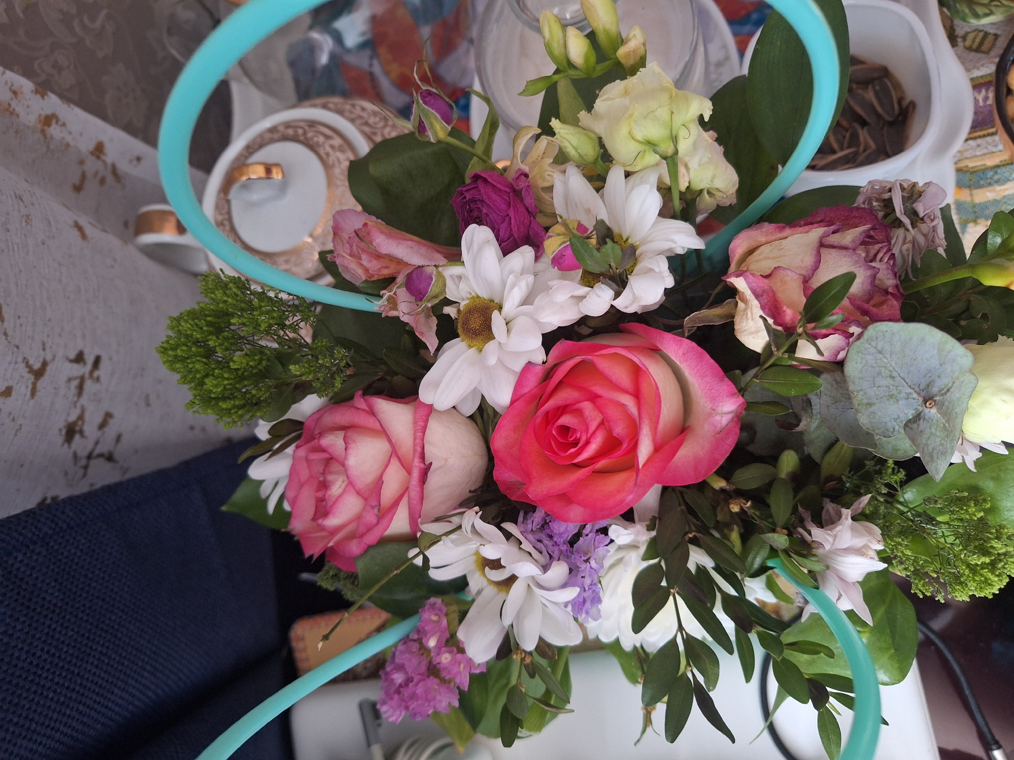A floral arrangement consisting of roses, daisies, and other flowers is displayed on a surface, possibly a table, against the backdrop of interior elements including a cup and saucer, a glass jar, and a container filled with seeds. The color scheme includes shades of pink, white, green, and purple. A turquoise circular band is placed around the flowers. The location cannot be determined.