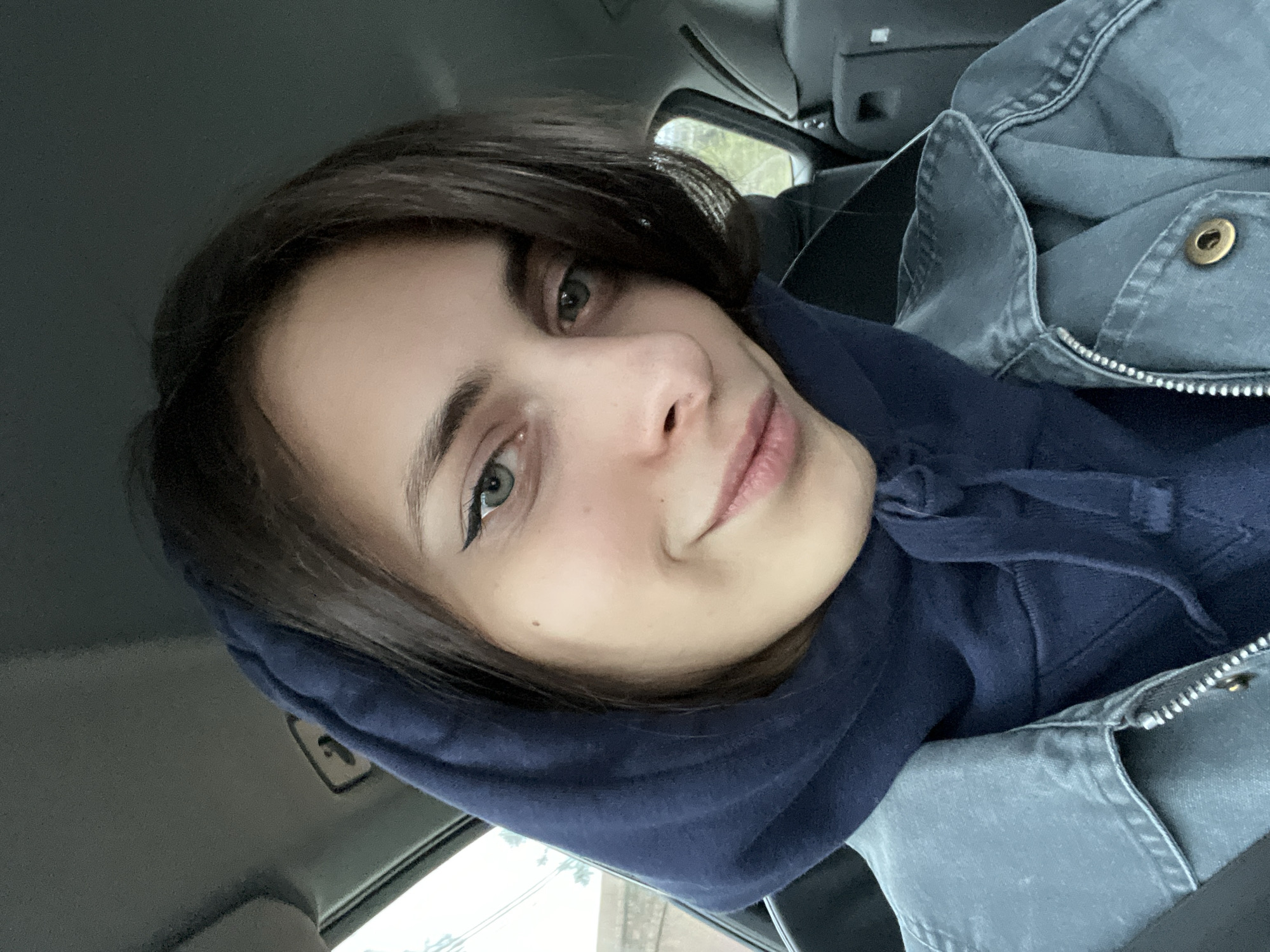 A young woman with short, dark hair and green eyes is wearing a dark blue hoodie and a denim jacket while sitting in a car. She is looking at the camera with a slight smile. The background shows a blurred street scene, suggesting the photo was taken during the day. Based on the presence of overhead powerlines and what appears to be a building exterior, the location could be a residential or commercial area. Without further visual cues or metadata, it is not possible to confirm the location in Krasnoyarsk, Russia.