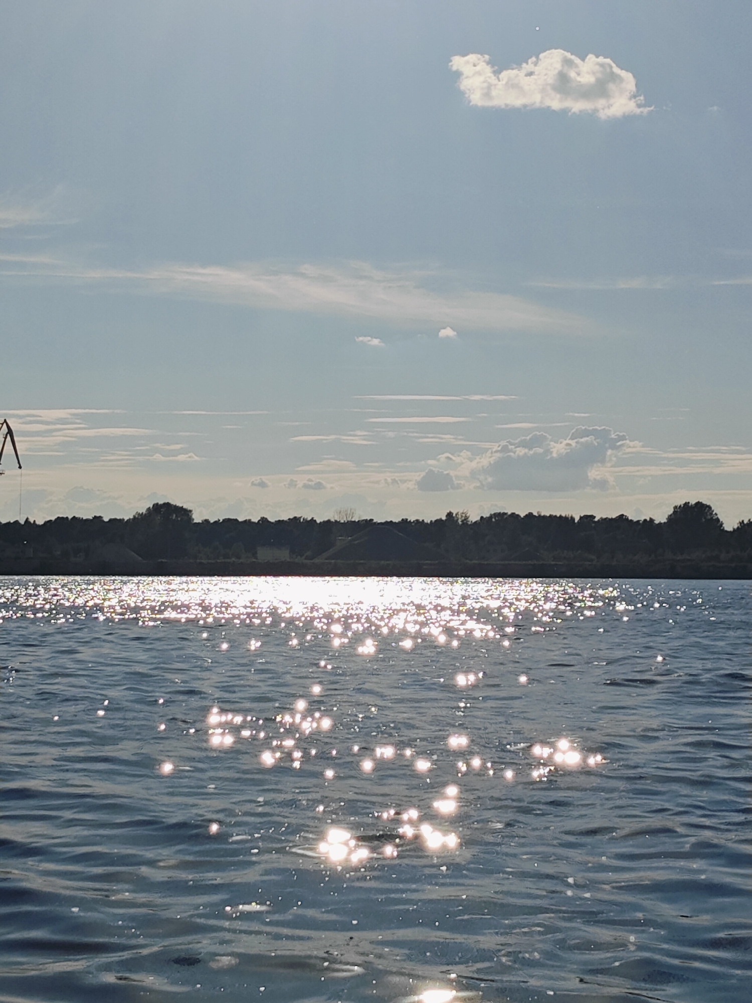 A wide-angle, slightly low-angle shot captures a sunlit body of water in Chaikovsky, Russia, with a treeline and a crane visible in the distance. The foreground is dominated by the rippling surface of the water, reflecting the sun in numerous bright, scattered glints. The water is a deep blue with darker wave crests and lighter troughs. Along the horizon, a dark, dense treeline forms a silhouetted barrier against the sky. To the left of the treeline, a tall industrial crane with a red and white striped boom is partially visible, its arm extending upwards. The sky above is a pale blue, with scattered white, wispy clouds. One larger, cumulus cloud is prominent in the upper right quadrant of the image. The lighting suggests it is daytime, likely late afternoon or early morning given the angle of the sun.