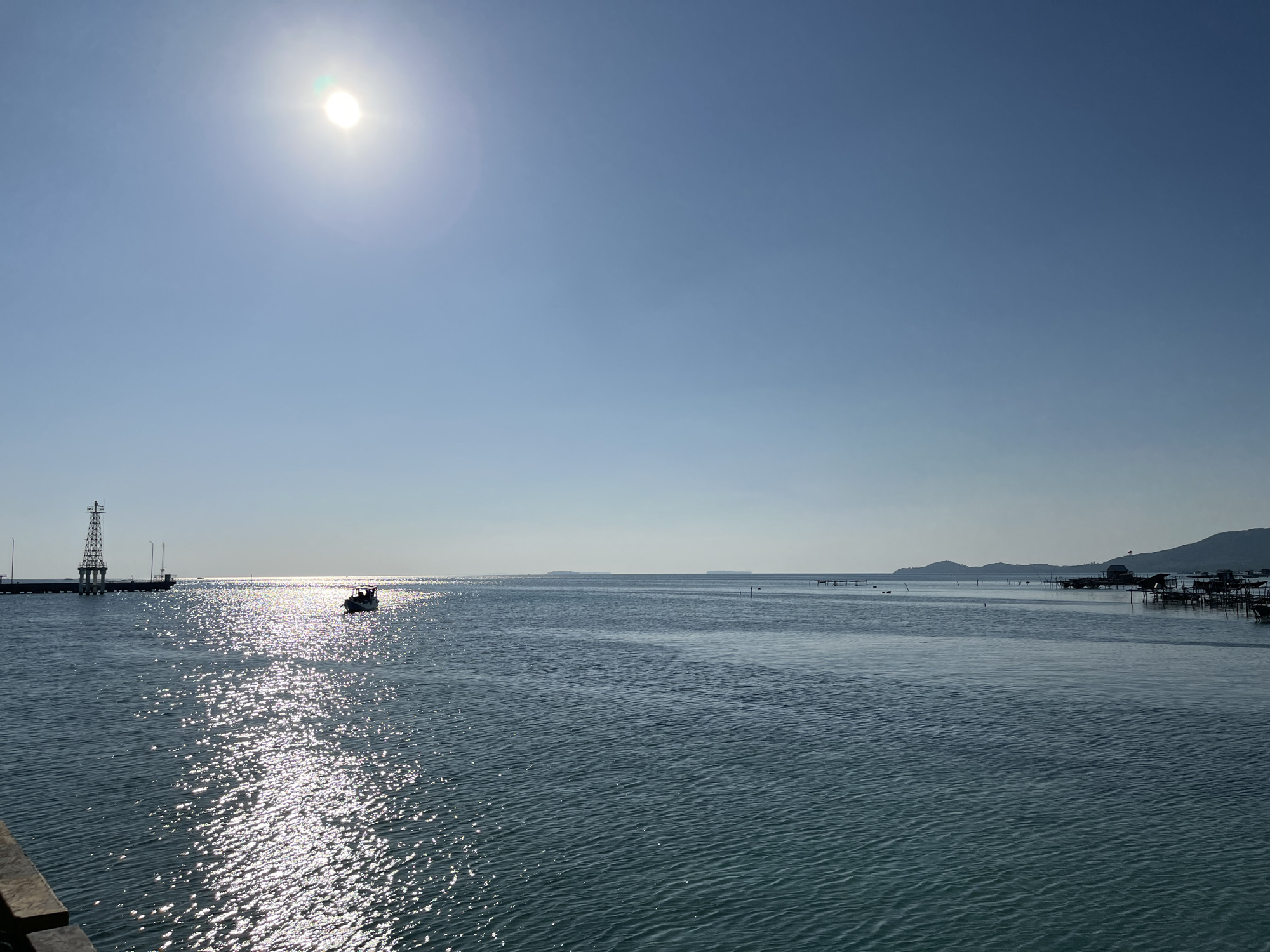 This is a wide shot of the ocean in Karimunjawa, Indonesia, taken during the day. The sun is high in the sky, casting a bright glare on the water. A small boat is visible in the middle of the frame, moving away from the viewer. To the left, a tall industrial-looking structure stands on a pier extending into the water. In the distance, hills or mountains can be seen on the horizon. The water is a deep blue-green, with gentle ripples across its surface. Some structures, possibly houses on stilts or fishing platforms, are visible on the right side of the frame, closer to the shore. The overall impression is of a clear, sunny day at sea.