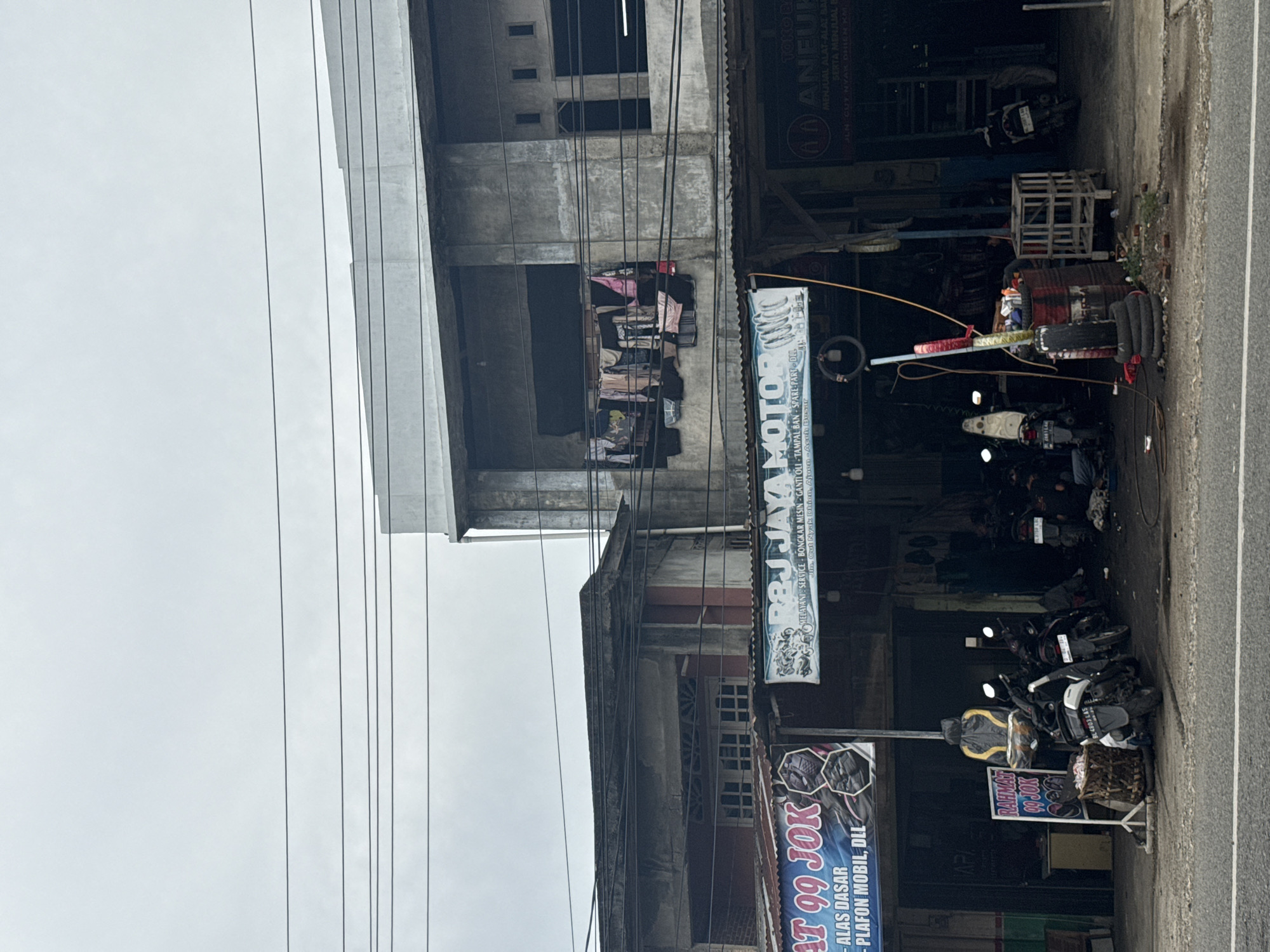 The image captures a street scene in Ajuen, Indonesia, with a focus on a collection of businesses lining the road. In the foreground, a paved road is visible, with a sidewalk or roadside area to its right. Several motorcycles are parked along the front of the buildings, suggesting a commercial area that caters to vehicle services or sales.
The buildings themselves appear to be multi-story structures with a mix of architectural styles. One building on the left features a corrugated metal roof and a banner with text, partially obscured, reading "AT 99 JOK - ALAS DASAR - PLAFON MOBIL, DLL". Next to it, a larger building displays a prominent sign for "R&J JAYA MOTOR", indicating a motorcycle repair shop or dealership. This sign also lists services like "MELAYANI: SERVICE - BONGKAR MESIN - GANTI OLI" and provides an address. Further to the right, another establishment, "ANEUK", is advertised with text suggesting it sells "ALAT-ALAT BAR" and "SERTA MENJUAL BA".
The upper floors of the buildings show varying states of upkeep. One section of a building on the right has several clothes hanging out to dry on a clothesline suspended across an open area. Electrical wires are strung across the scene, typical of urban or semi-urban environments in Indonesia. The sky is overcast, contributing to a diffused, natural light across the entire image. The overall impression is that of a busy, functional commercial district.