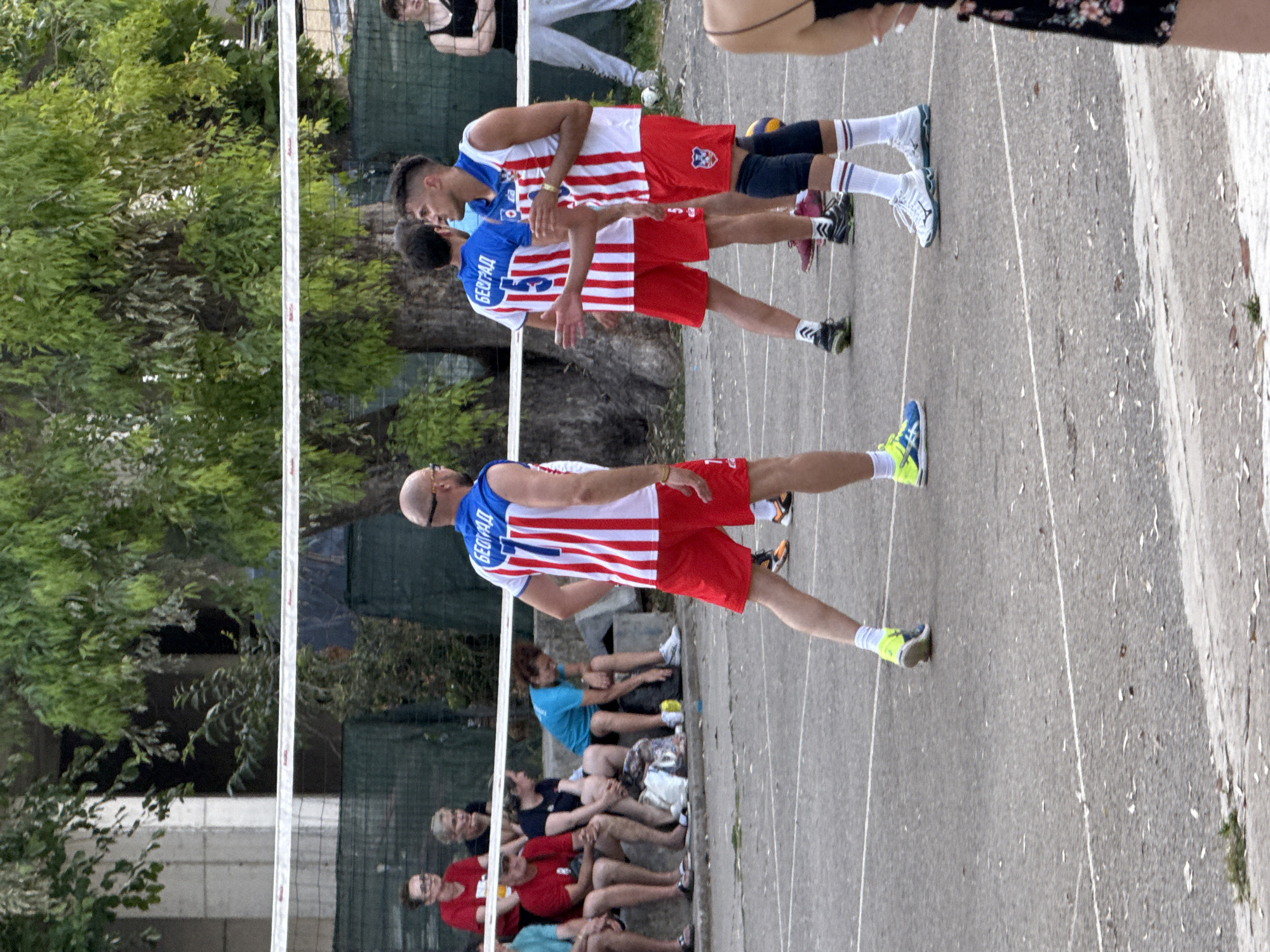The image depicts an outdoor volleyball game in Budva, Montenegro. Three male volleyball players, dressed in red and white striped jerseys and red shorts, are on the court. One player in the foreground faces away from the camera, looking towards the net. Another player, in the middle ground, is turned to his right, his left arm extended as if in mid-conversation or instruction. A third player, further back on the right, is also looking to his right, his right arm raised slightly. Several spectators are seated on a concrete bench behind the net, some in red shirts, others in dark tops. The court appears to be made of asphalt, with white lines marking its boundaries. Green foliage and trees form a backdrop, with a concrete building visible behind the trees. The overall scene is informal, suggesting a recreational or amateur match.