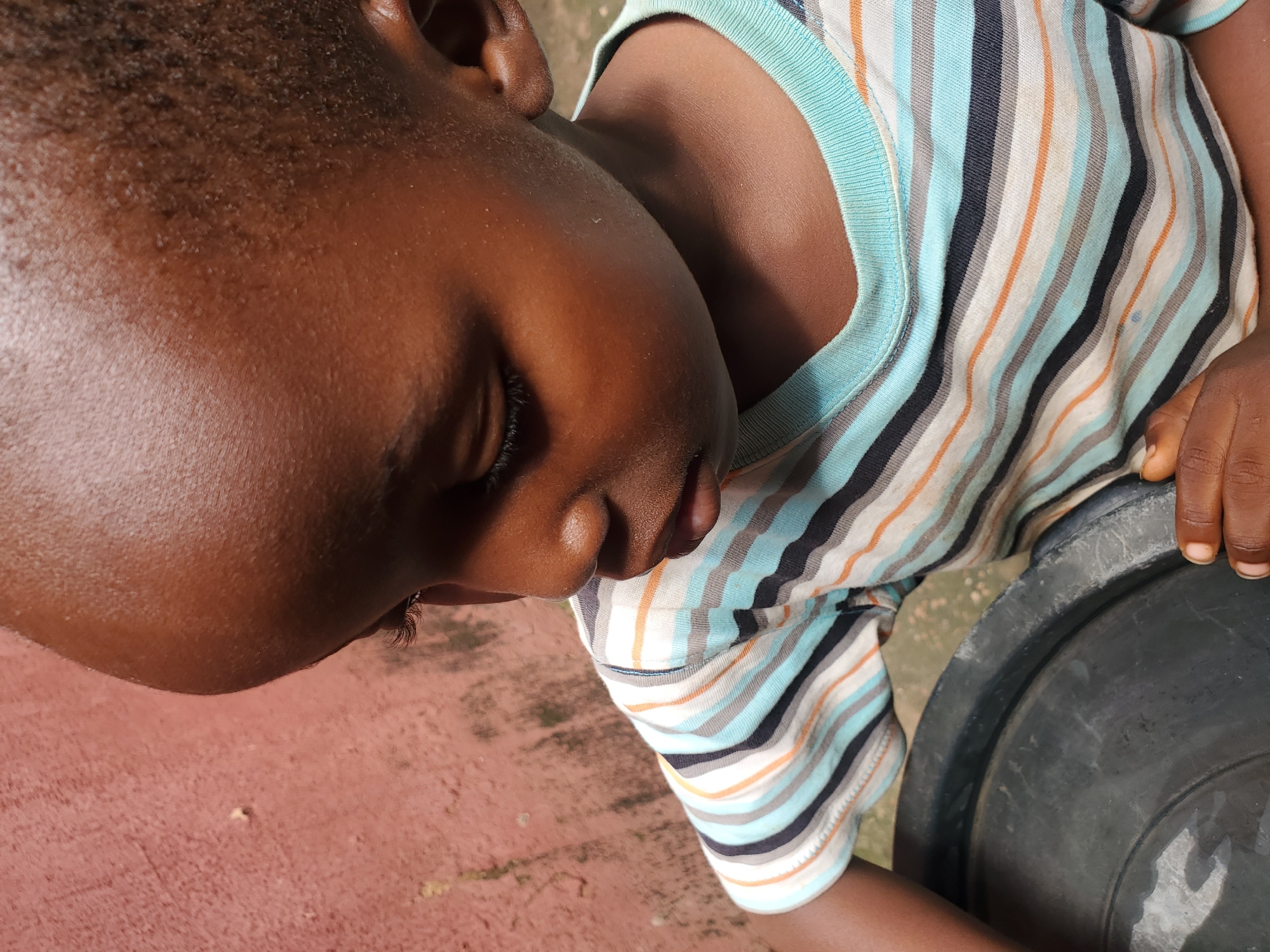 This media file is a close-up, high-angle shot of a young child in Jos, Nigeria. The child is lying down on a reddish, textured surface, possibly an outdoor ground. Their face is turned to the left, with their eyes closed, suggesting they are sleeping or resting. The child has dark skin and short, dark hair. They are wearing a horizontally striped shirt with alternating bands of blue, white, black, and orange. The child's left arm is bent, and their hand is resting near a large, dark, circular object, which appears to be a plastic basin or bucket. The lighting suggests it is daytime, with soft shadows indicating the sun is not directly overhead. The overall impression is one of peaceful rest, possibly in an informal outdoor setting typical of a residential area in Nigeria. There is no visible text in the image.