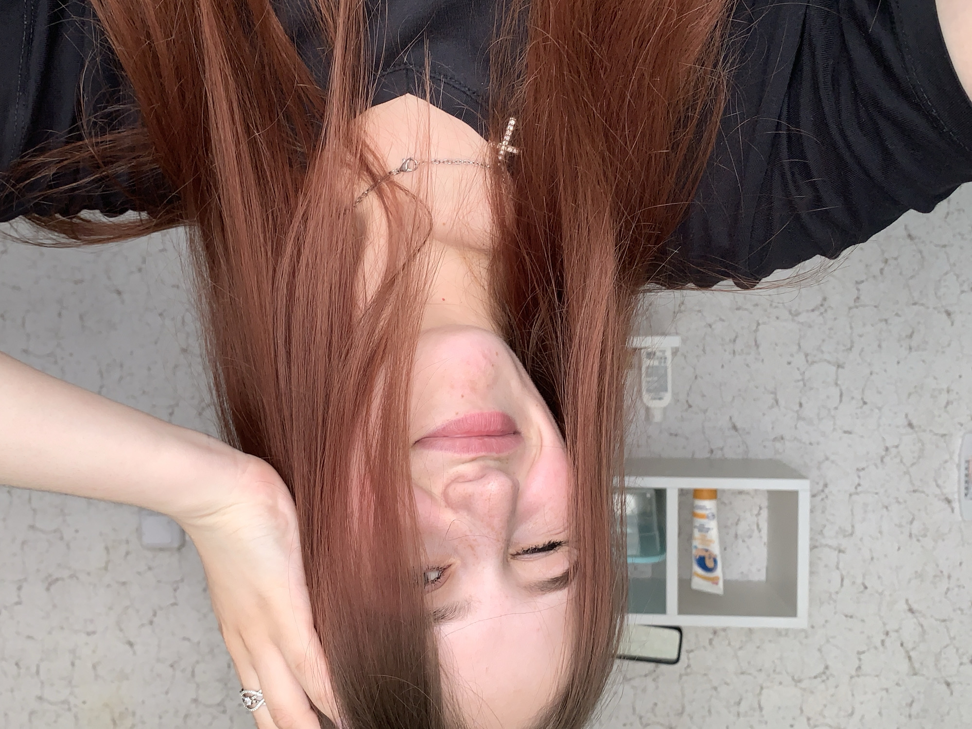 A young woman with long, auburn hair and fair skin is pictured upside down. She wears a black shirt and a silver necklace with a cross pendant. Her left hand, adorned with a silver ring, supports her head. The background features a white shelf with a blue water bottle and a tube of sunscreen, and a wall with a subtle, patterned design. The location is Фоки, Russia.