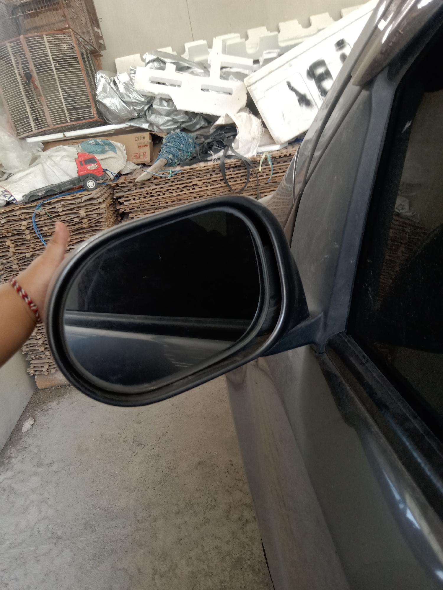 A hand with a braided red, white, and black bracelet is reaching out towards a car's side-view mirror. The mirror is black and oval-shaped, reflecting the dark interior of the car and a blurred exterior. The car appears to be parked in an outdoor area with a concrete floor. In the background, there is a stack of corrugated cardboard, a white styrofoam object with black markings, a silver-colored tarp, and a red toy truck. The scene is likely in Stiaji, Indonesia, given the context.