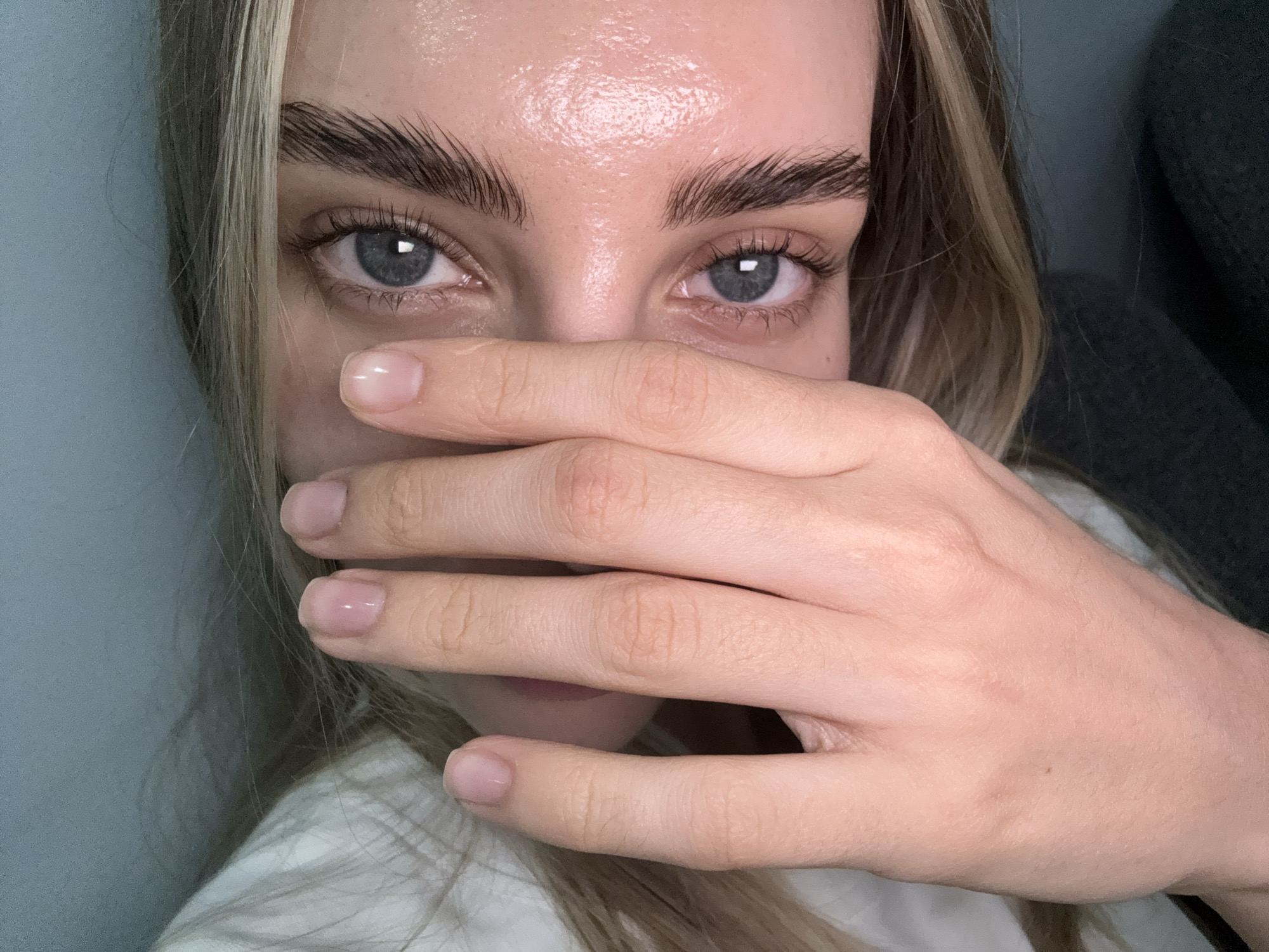The image is a close-up, indoor shot of a young woman covering her mouth and nose with her hand. Her eyes, with blue irises and dark lashes, are visible, as are her eyebrows, which appear to be brushed upwards. Her skin is smooth and has a dewy appearance. Her fingernails are short and polished with a sheer pink shade. Her blonde hair is visible on the left side of the frame, framing her face. The background is a plain, light blue wall on the left and a dark, textured surface on the right, possibly a piece of furniture or clothing. The lighting is bright, highlighting the texture of her skin. There are no discernible elements in the image that would indicate a specific location in Stavropol, Russia.