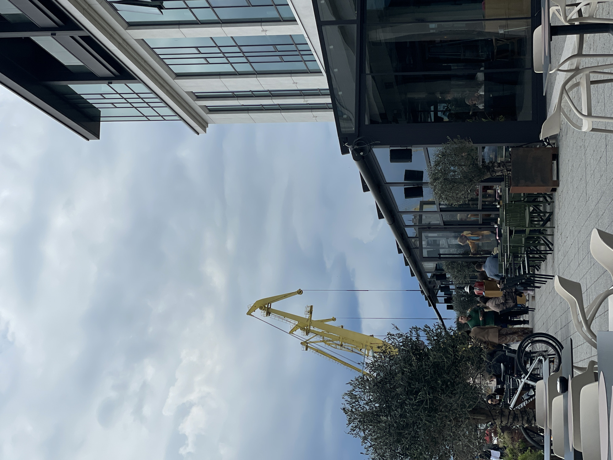 The image depicts a street scene in Burgas, Bulgaria. The sky is overcast with light clouds. Two modern buildings frame the upper edges of the view. One building has a glass facade with visible window gridlines, while the other has a more traditional architectural style. A large yellow crane is positioned prominently in the middle ground, partially obscured by a tree.
In the foreground, there is a restaurant or cafe with outdoor seating. People are seated at tables. Bicycle parked next to the eating area. White plastic chairs are arranged around tables. Reflections of people can be seen in the glass facade of the restaurant.