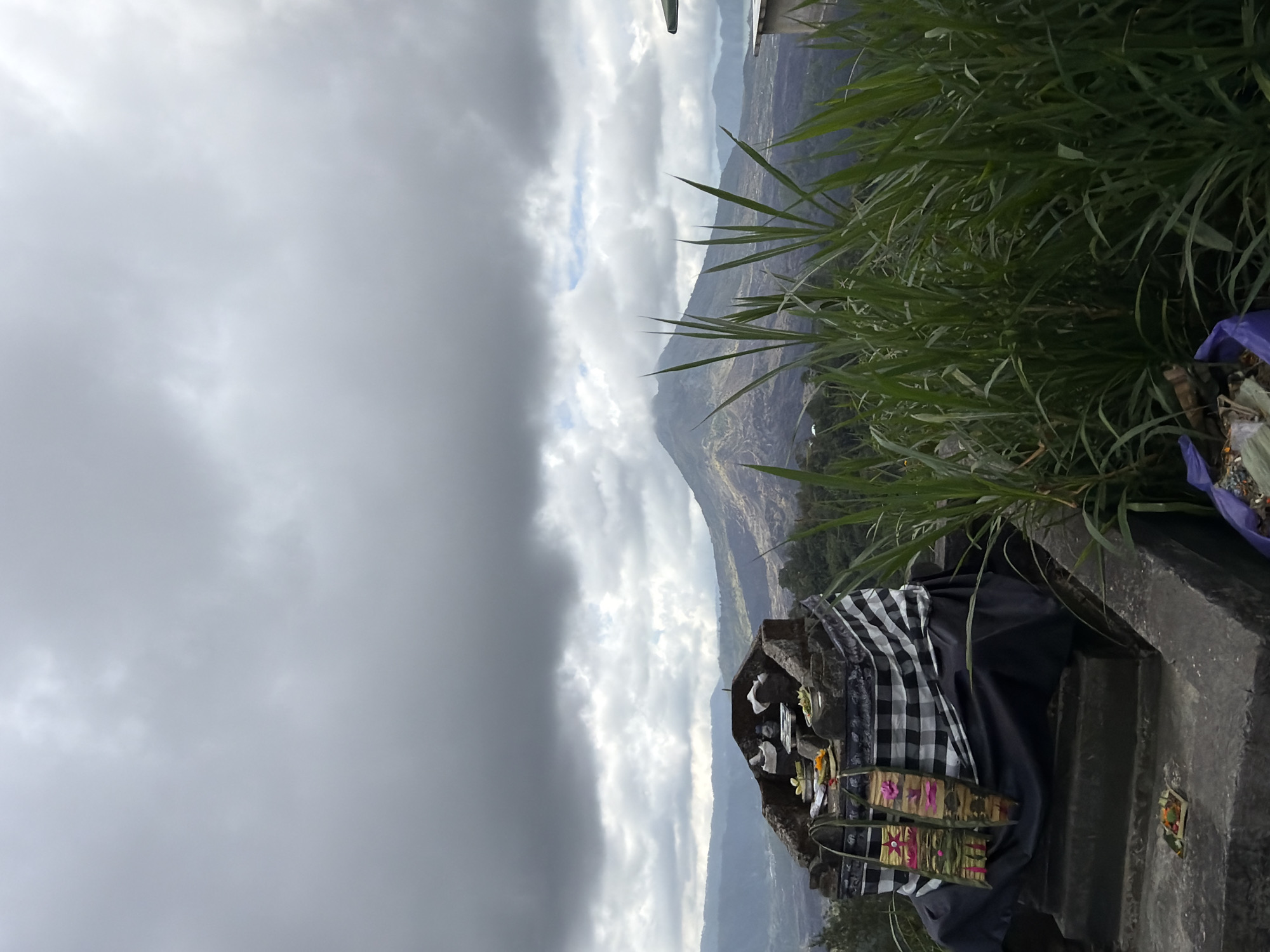 The image depicts a scenic overlook in Kintamani, Indonesia, with a view of Mount Batur. In the foreground, there is a stone altar adorned with offerings. The altar is draped with a black cloth and a black and white checkered fabric. Two decorative banners, intricately woven with pink floral patterns, hang from the altar. Small woven baskets containing flowers and other offerings are placed on the altar and on a lower stone ledge. Lush green grass and reeds are in the immediate foreground, partially obscuring the view. In the background, Mount Batur rises majestically, its slopes covered in dark, volcanic rock. The sky is overcast with heavy gray clouds, casting a dramatic mood over the landscape. A body of water, likely Lake Batur, is visible in the distance, reflecting the muted light. The overall scene captures a spiritual and natural landscape characteristic of the region.