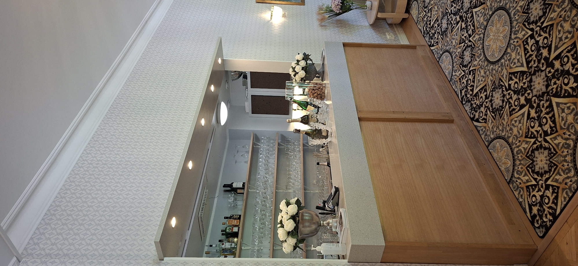 This is a medium shot, eye-level, indoor photograph of a bar area, likely in Lahti, Finland. The bar itself is constructed with a light-colored speckled countertop and a wooden base, featuring sliding panel doors. Behind the bar, shelves are stocked with numerous wine glasses, champagne flutes, and various bottles of alcohol. Two bottles of champagne are chilled in ice buckets on the countertop, alongside a vase of white roses and a jar of nuts. A hand sanitizer dispenser is also visible on the left side of the counter. The walls are covered in a patterned wallpaper. The floor is carpeted with a dark rug featuring a complex, repeating geometric pattern in gold and grey. A circular side table with a vase of pink flowers is visible to the right. No people are present in the image.