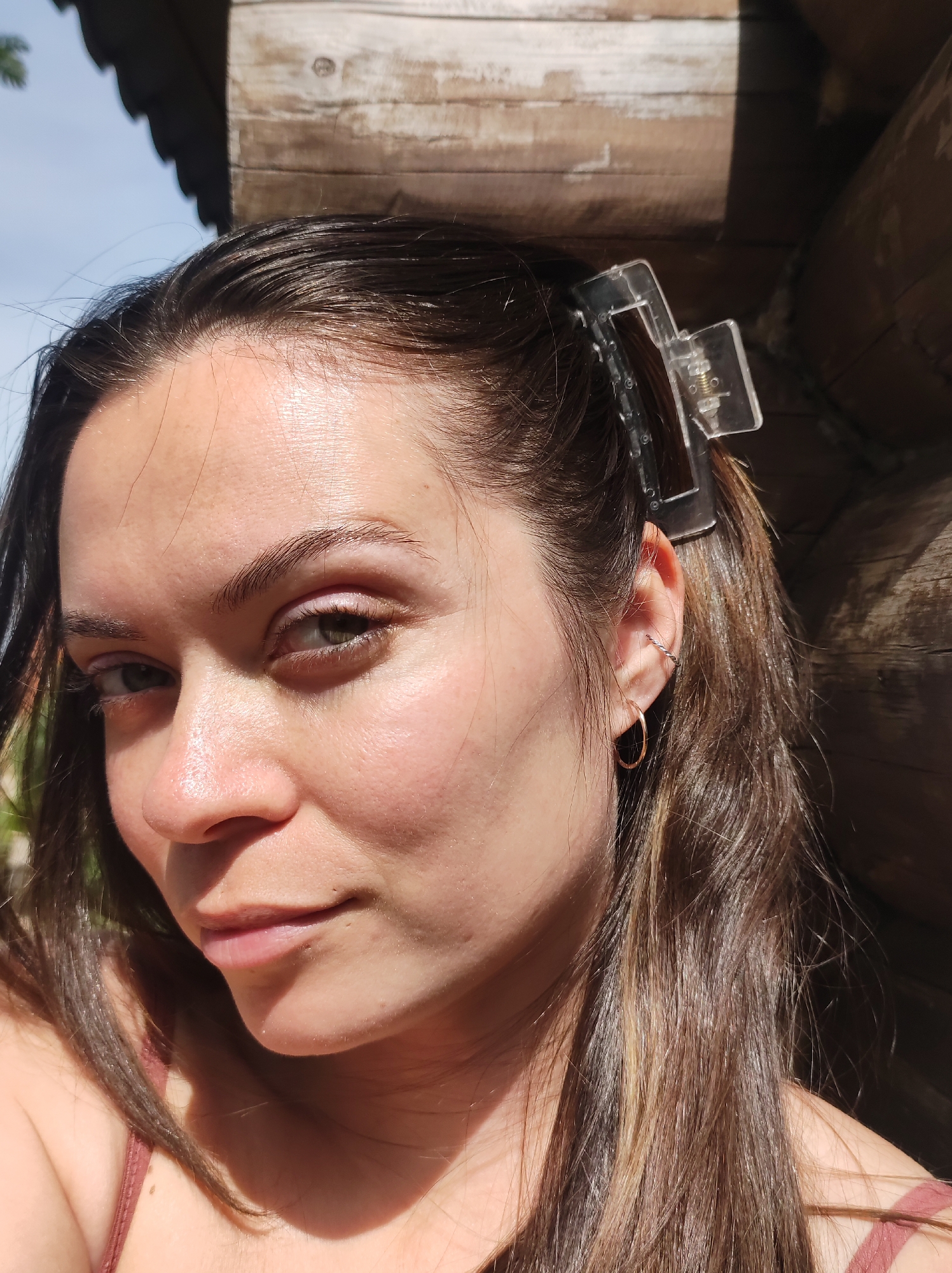 A close-up shot of a woman with brown hair, some of which is pulled back with a clear hair clip, standing next to a log structure. She has light skin, brown eyes, and is wearing earrings. The background is out of focus, but the log structure is visible, suggesting a possibly rural or wooded setting. Sunlight is casting shadows on her face and the structure. The location in Фоки, Russia is not discernible from the image alone.