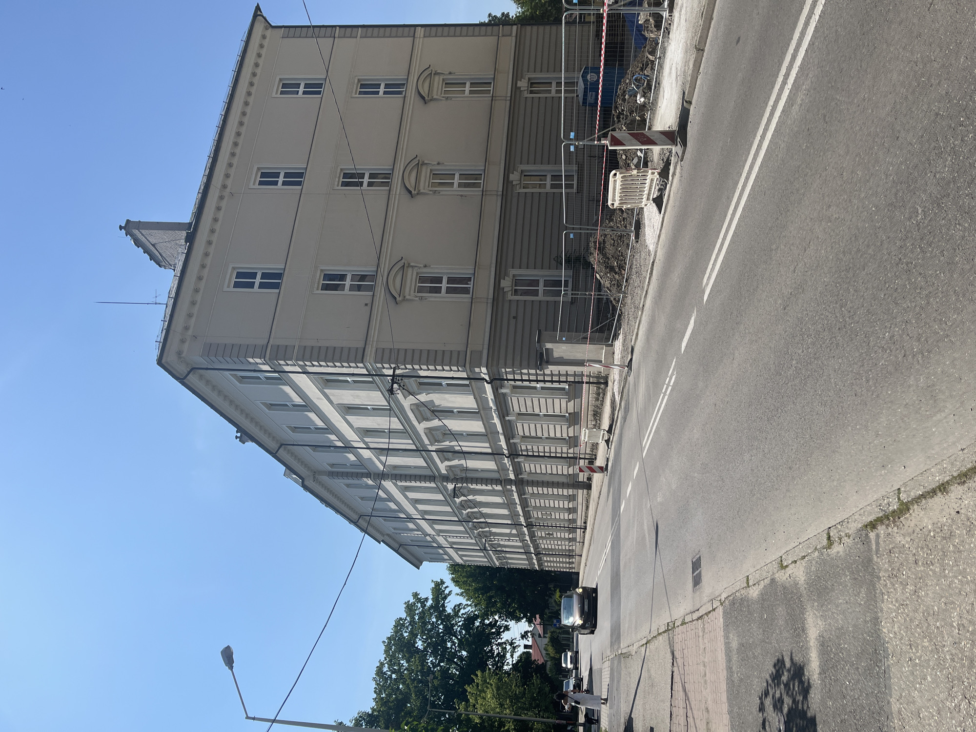 The image shows a street in an urban environment under a clear blue sky. A large, pale yellow, multi-story building with numerous windows lines the right side of the street. The building has a classical architectural style with decorative elements along the roofline and around the windows. Construction fencing with red and white tape is visible along the base of the building, indicating ongoing work. A paved road with white lane markings runs through the center of the image. Several cars are present on the road, with one in the foreground appearing to be dark colored. A woman in a white dress is walking on the sidewalk on the left side of the street, accompanied by a dog. Trees with green foliage are present on the far left side of the image, and a streetlamp is also visible. The ground appears to be a mix of paved sidewalk, asphalt, and dirt where the construction is taking place.