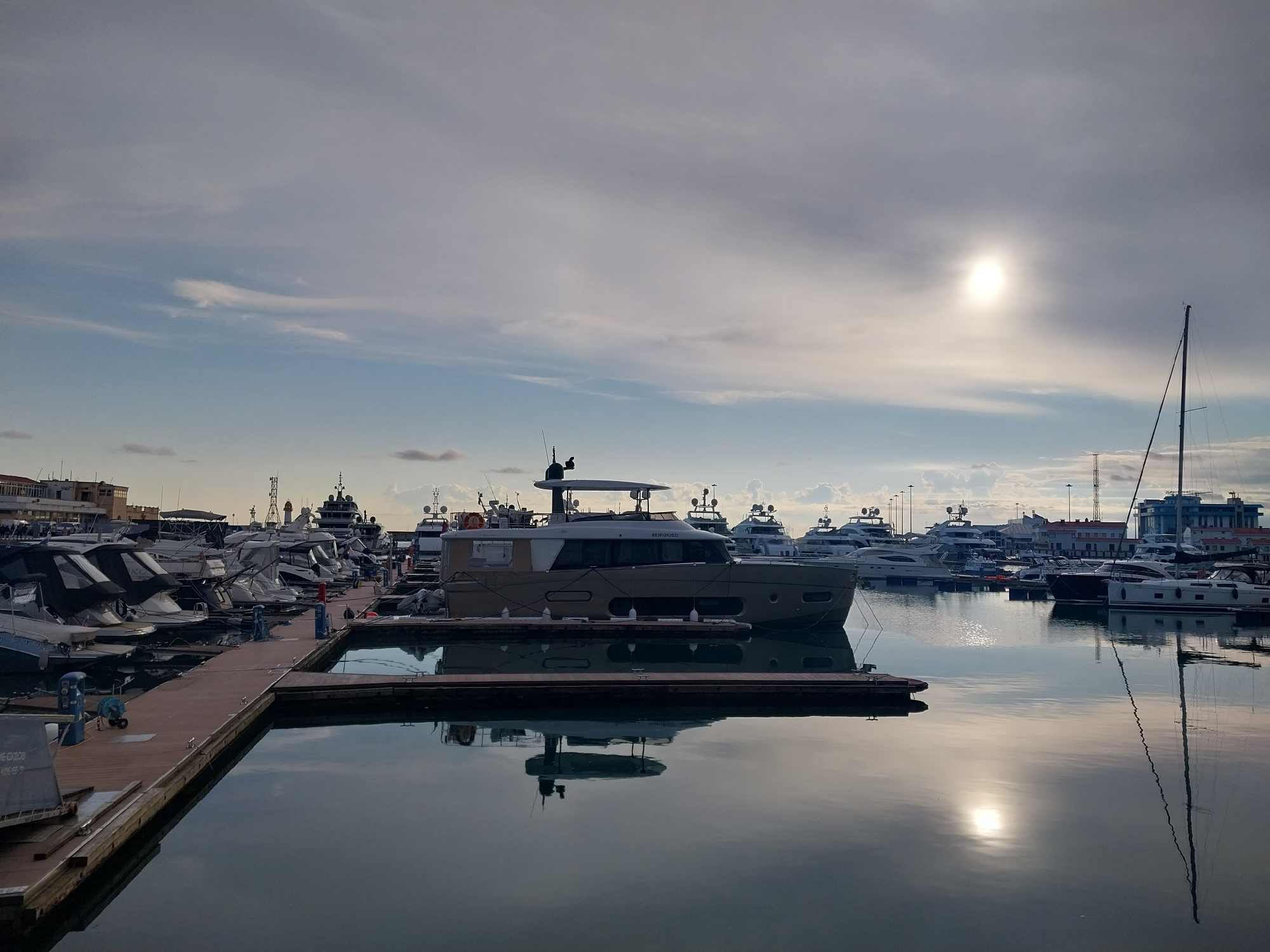 The image depicts a marina in Sochi, Russia. A modern yacht is docked at a floating pier in the foreground, its reflection visible in the still water. Numerous other boats, including sailboats and motor yachts, are moored in the background, creating a crowded harbor scene. A breakwater or pier extends into the water on the left side of the frame. In the background, buildings and structures of the city are visible, including communication towers and buildings. The sky is overcast with the sun visible.