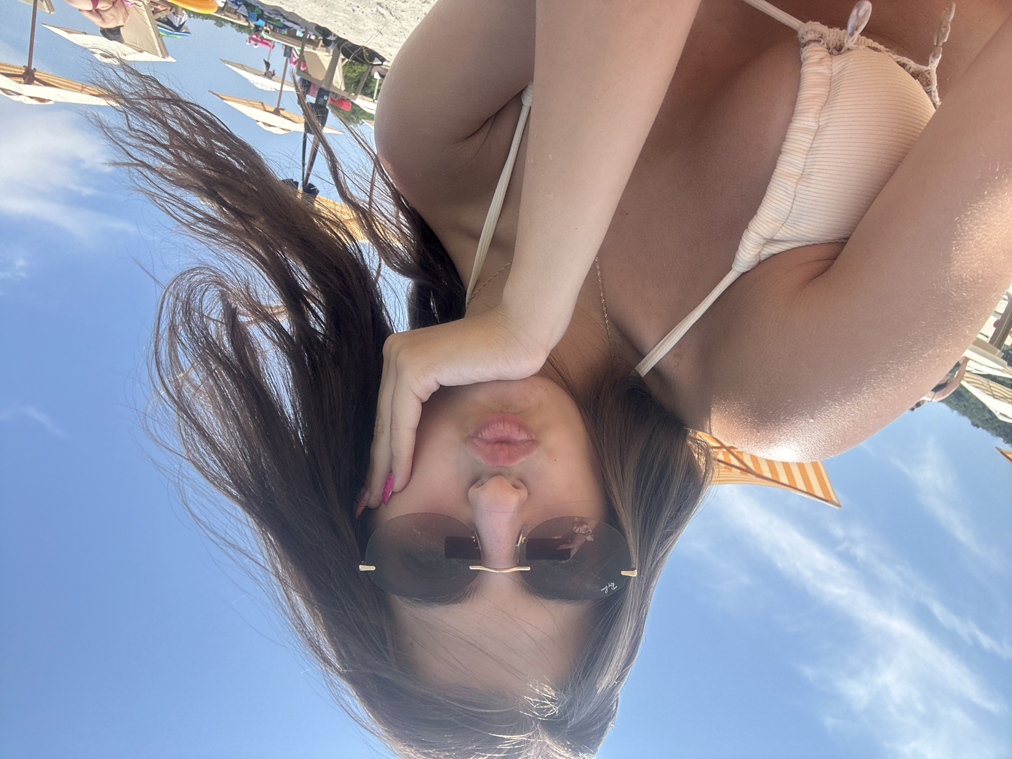A young woman with long, dark hair is taking a selfie on a sunny day at a beach in Varna, Bulgaria. She is wearing a light-colored bikini and sunglasses. Her hand is resting on her chin, and her lips are pursed as if she is blowing a kiss. In the background, there are beach umbrellas, lounge chairs, and other beachgoers. The sky is blue with a few scattered clouds.