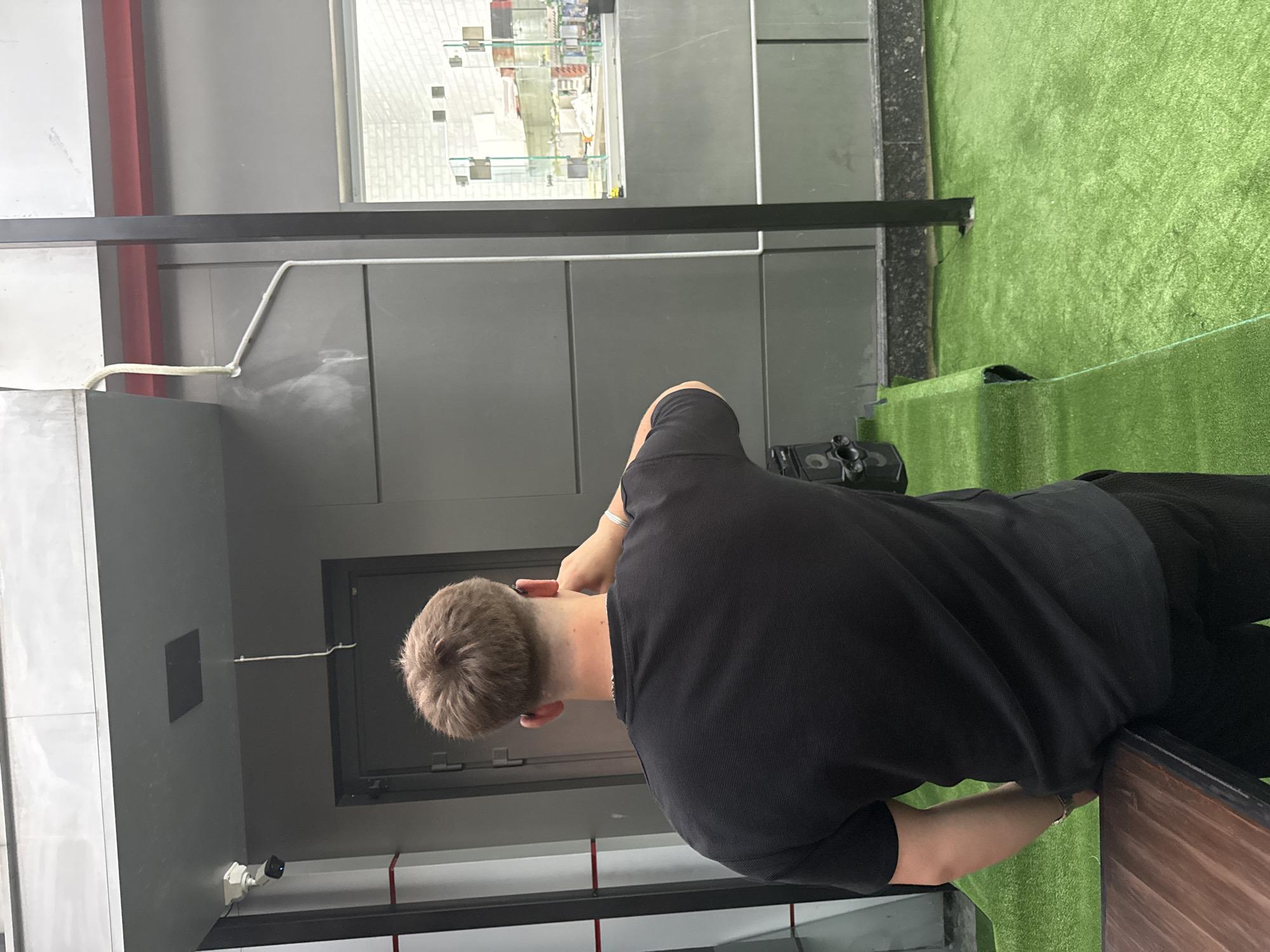 The image shows a person from behind, wearing a black t-shirt and black pants, standing on bright green artificial turf. The person has a light brown, short haircut. To the left of the person, there is a security camera mounted on a white pole. Behind the person is a dark gray building facade with a closed door. Above and to the right of the door, a white pipe extends from the building, with a visible mist or steam emanating from it. Further to the right, there is a large window with a view of what appears to be a store interior with shelves stocked with various items. A black speaker is visible on the turf to the left of the person. A wooden table edge is in the lower-left corner of the image. The overall setting appears to be an outdoor area, possibly a patio or a commercial space, in Chișinău, Moldova.