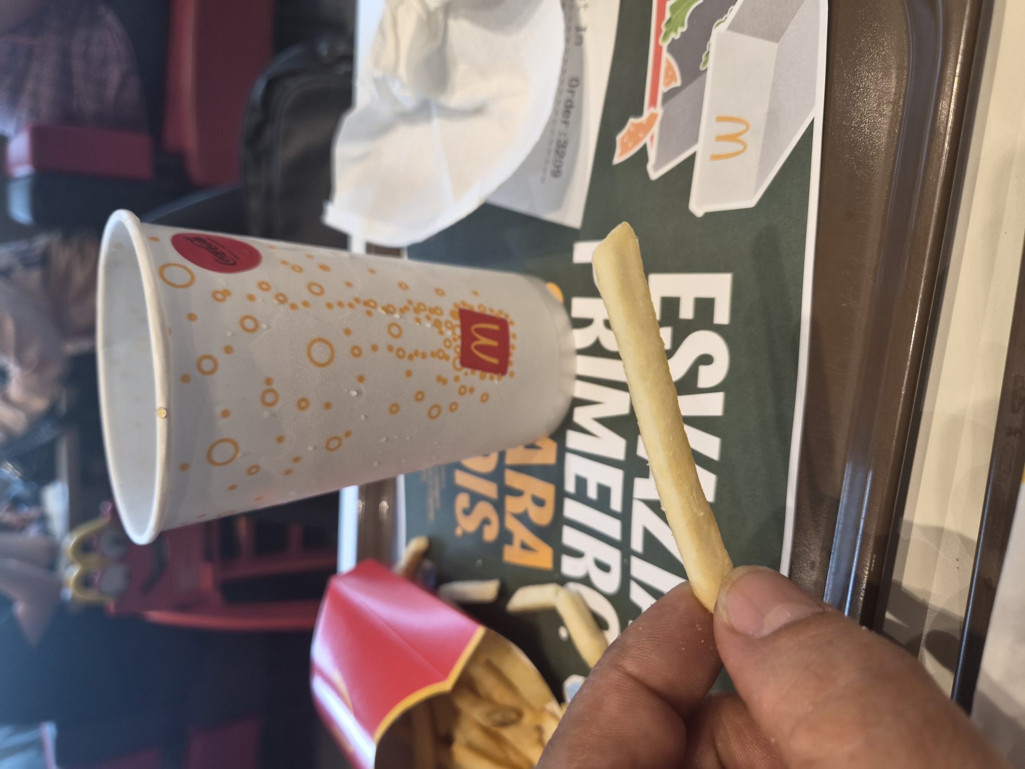 The image shows a close-up of a hand holding a french fry over a tray. In the background, there is a McDonald's cup with a Coca-Cola logo and a red McDonald's fry box. The tray has some text and graphics, including what appears to be an order number. The scene is likely at a McDonald's restaurant, possibly in Bragança, Portugal, given the context of the query. The overall lighting suggests an indoor setting.